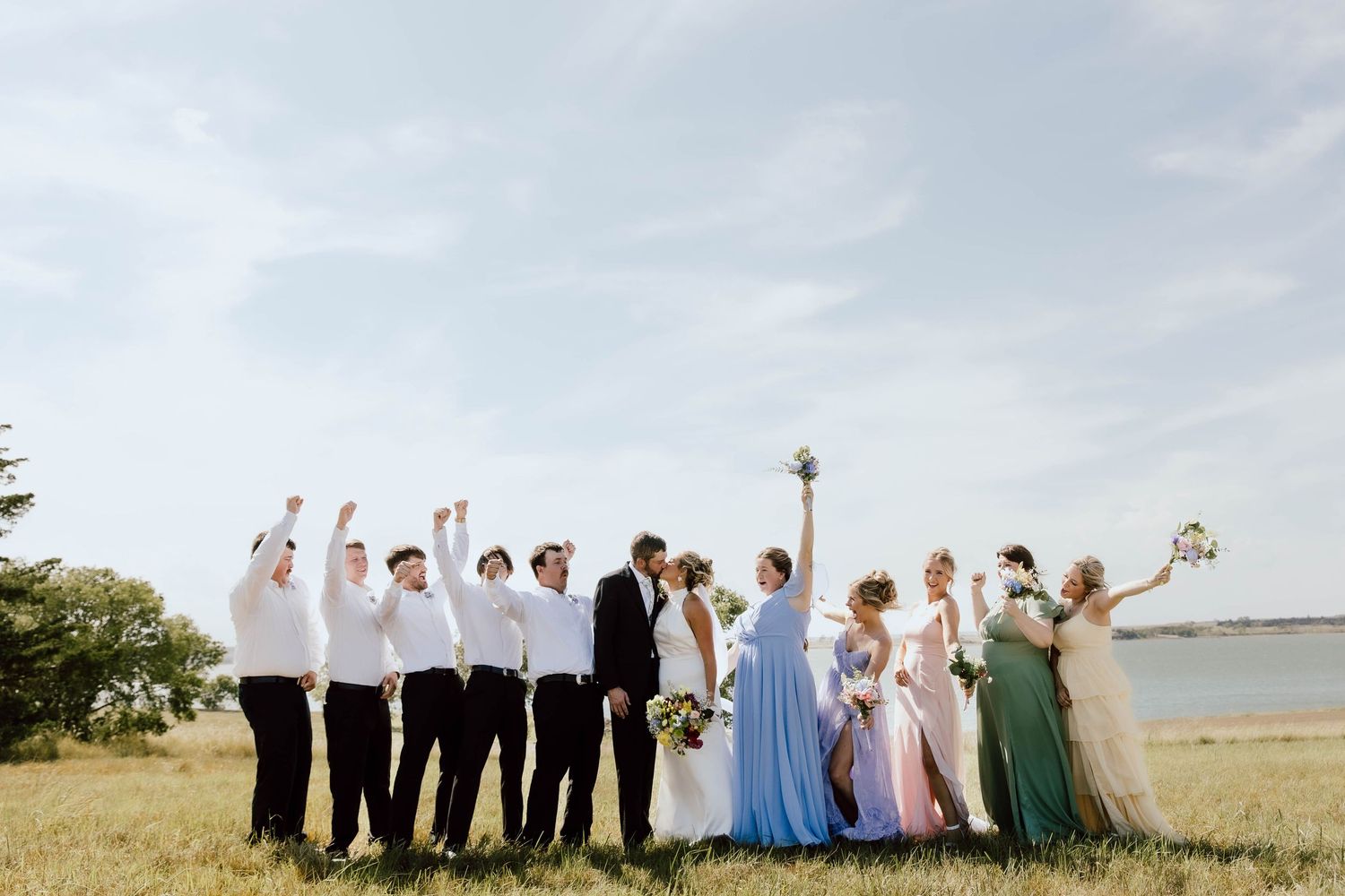 A wedding party poses together in a field with their hands raised joyfully against a cloudy sky.