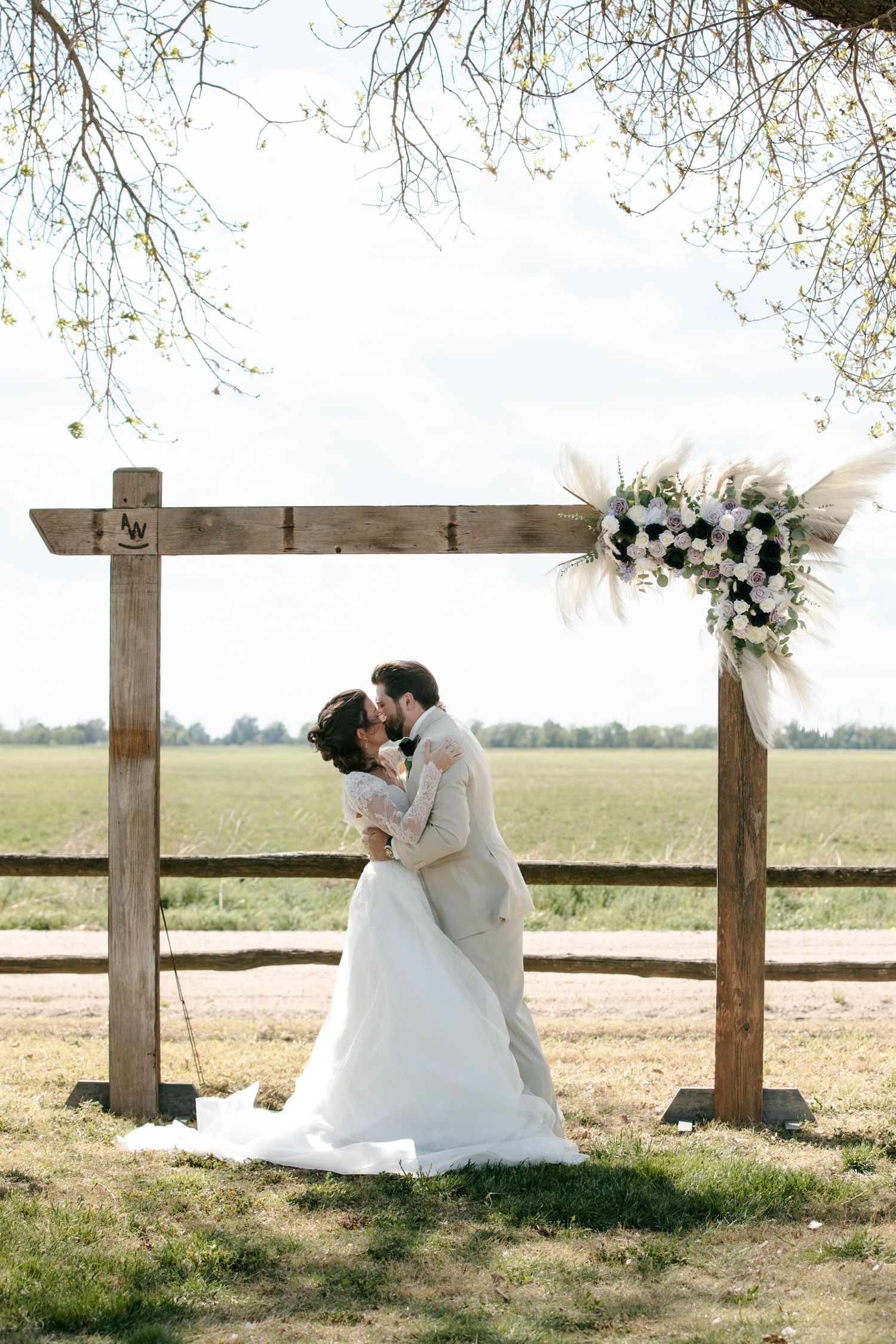 Bride and groom share a kiss under a rustic wooden arch adorned with white floral arrangement in an outdoor field setting.