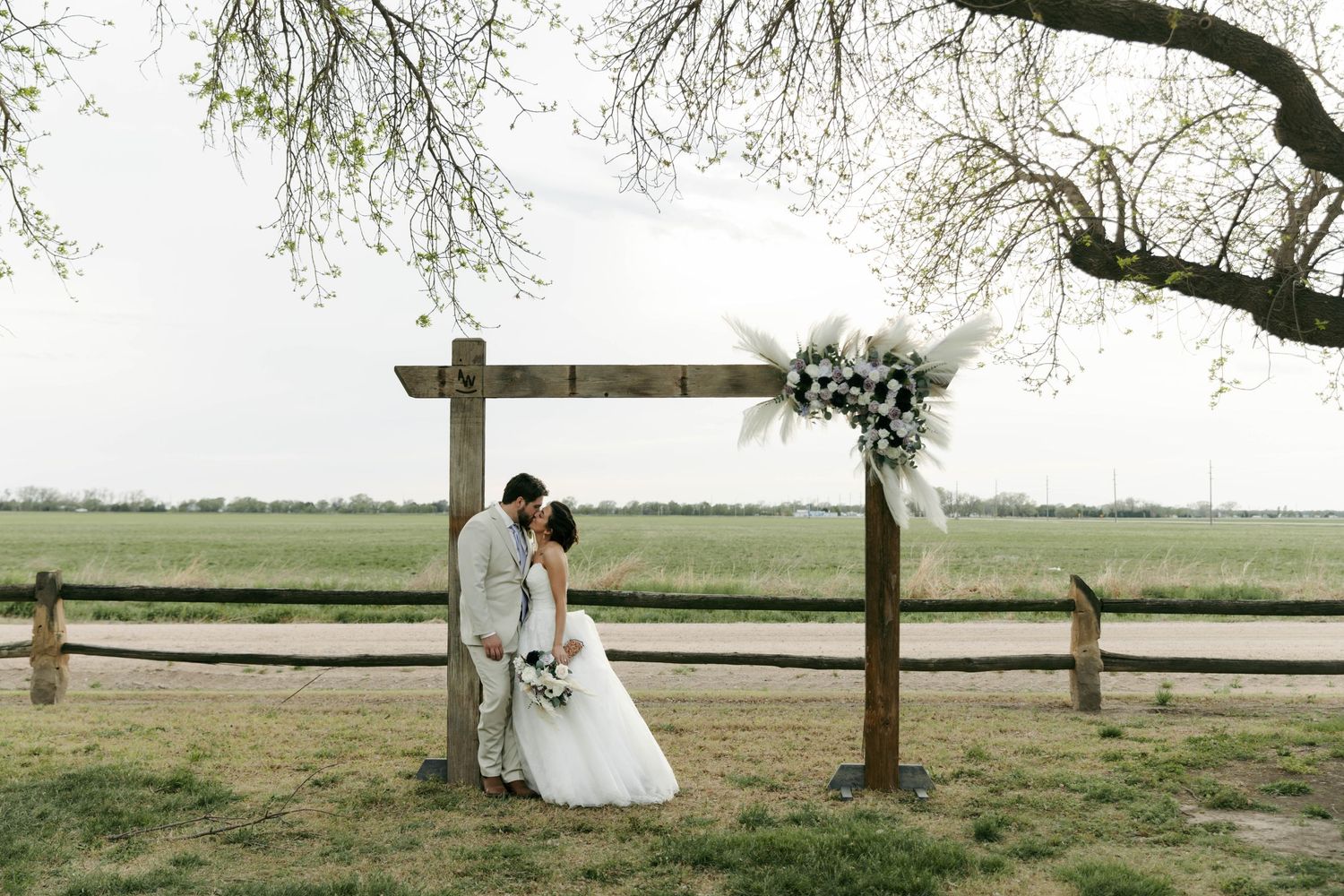 Bride and groom kissing under rustic wooden arch with floral wreath in an open green field.