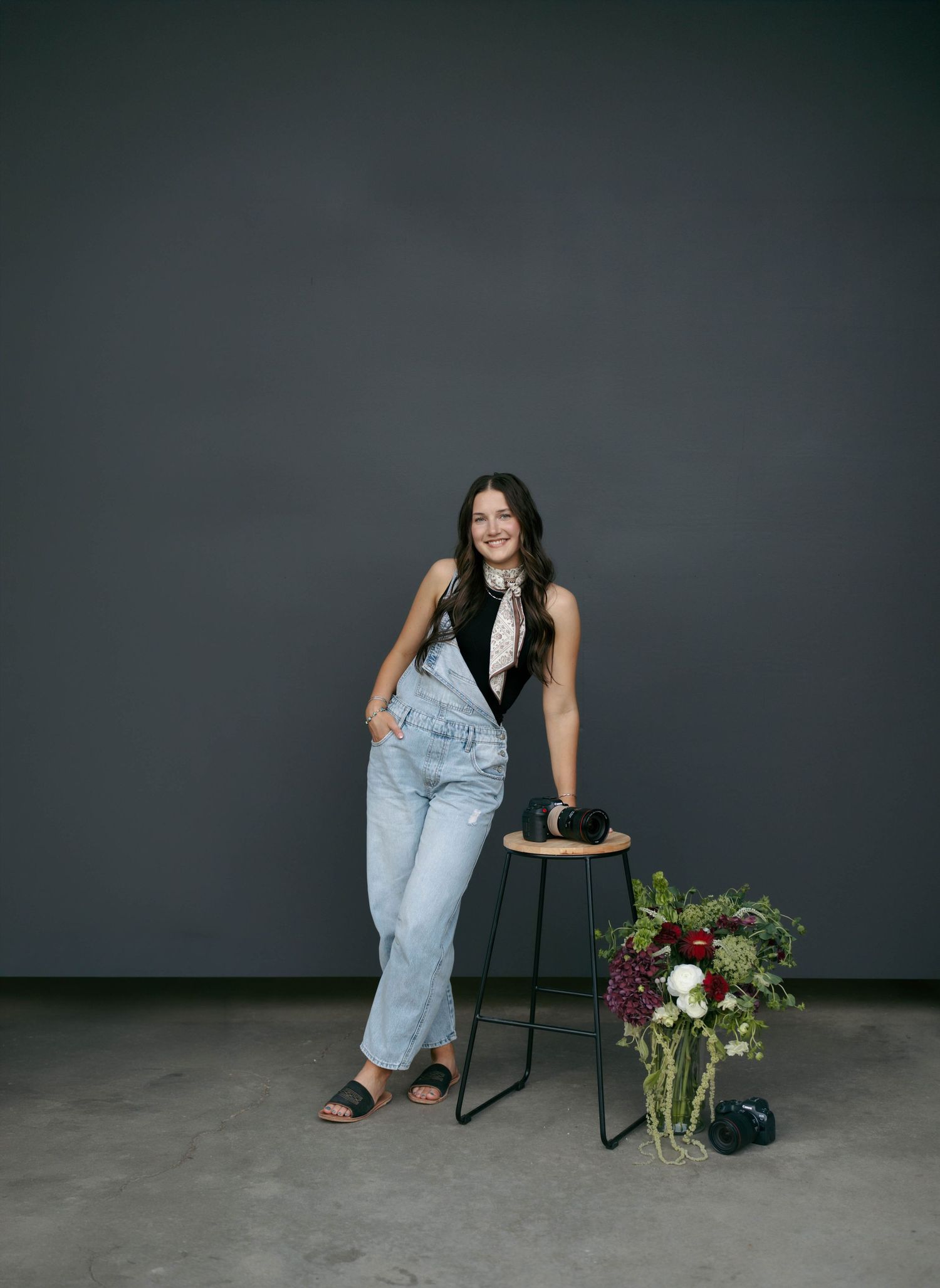 Woman in denim overalls smiling beside a floral arrangement on a stool against a dark gray backdrop.