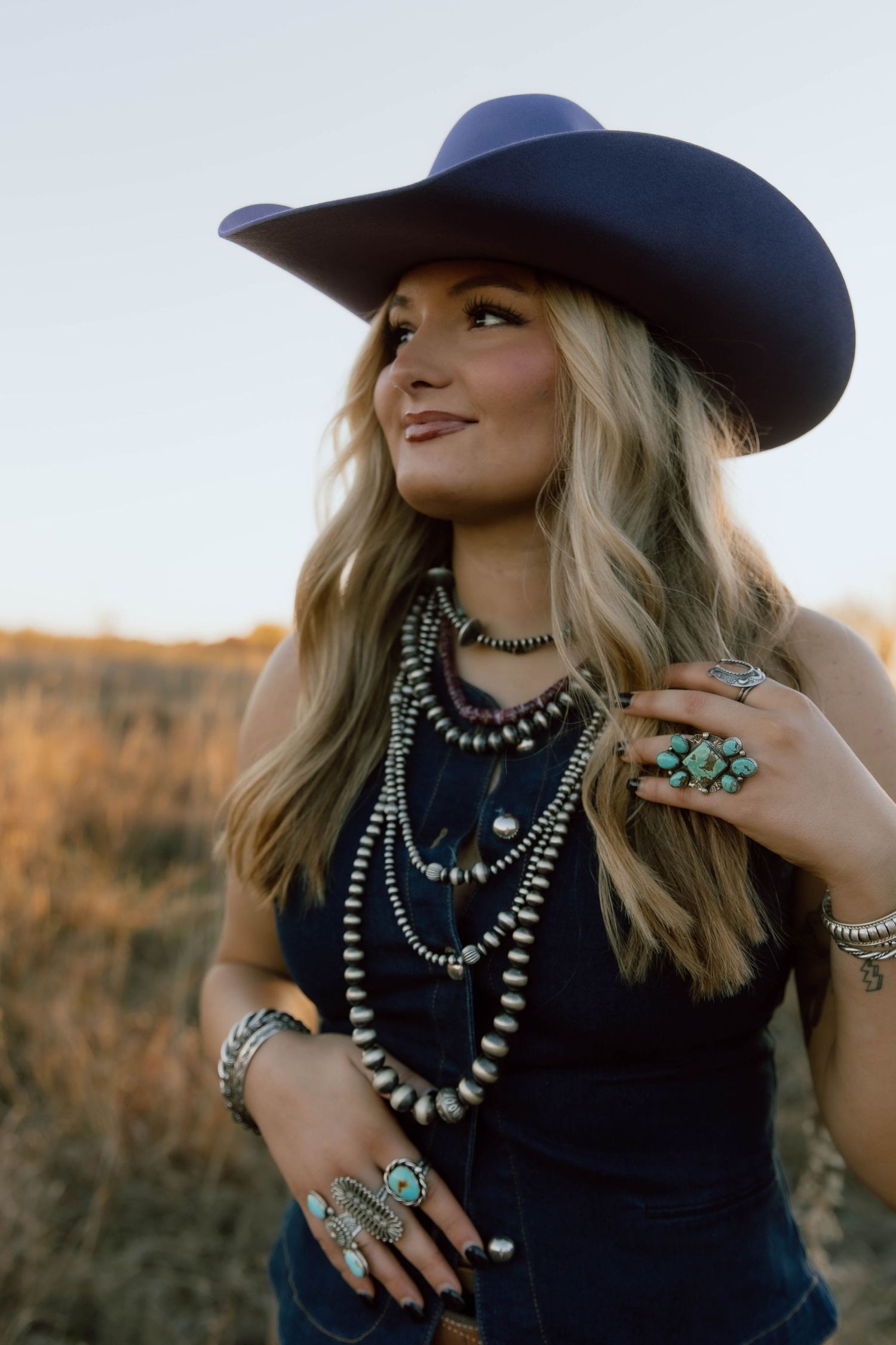 Western style portrait with navy cowboy hat and layered silver necklaces at sunset.