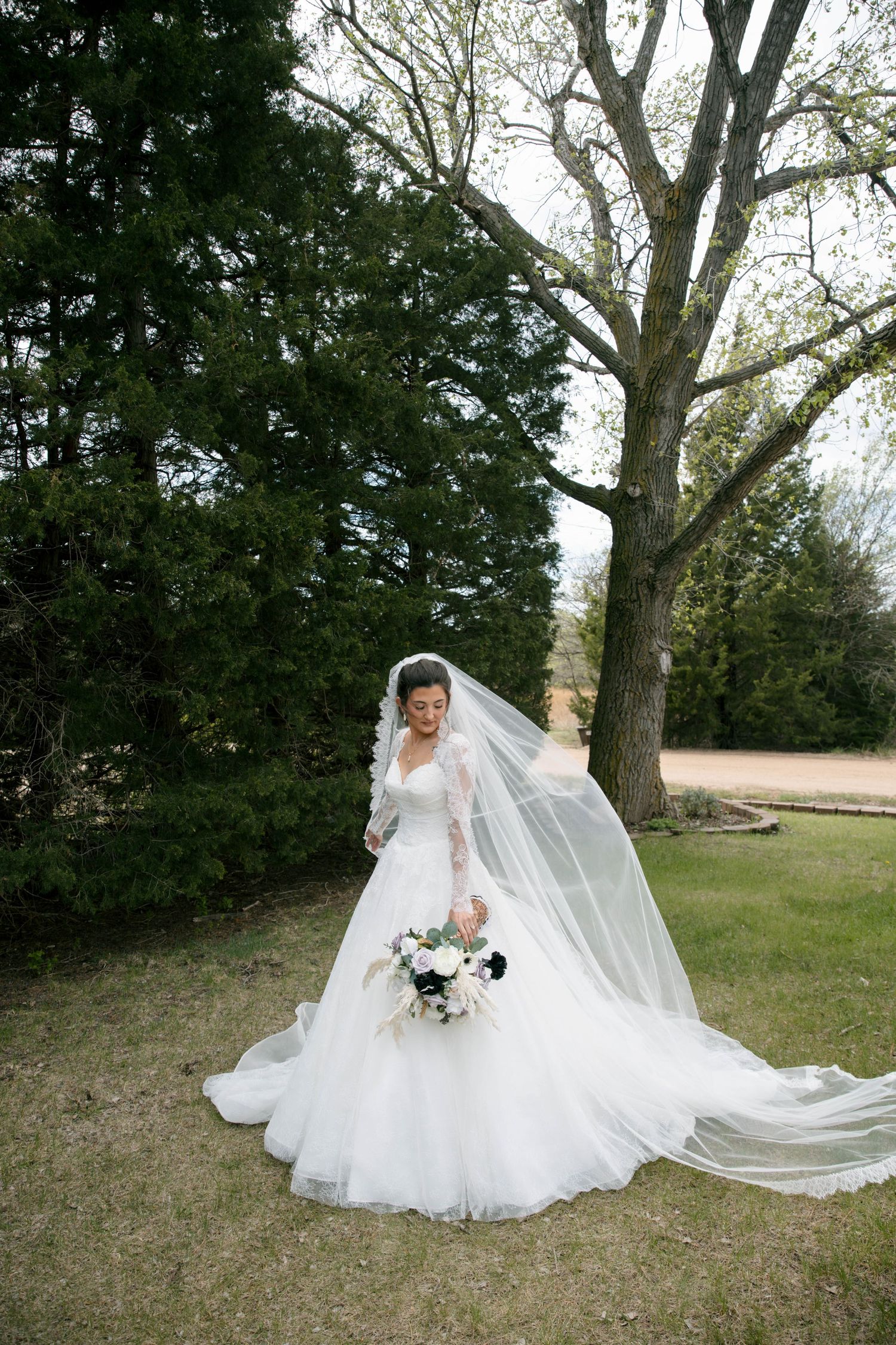 Bride in a white ball gown with long veil holding a bouquet, standing outdoors among lush green trees.