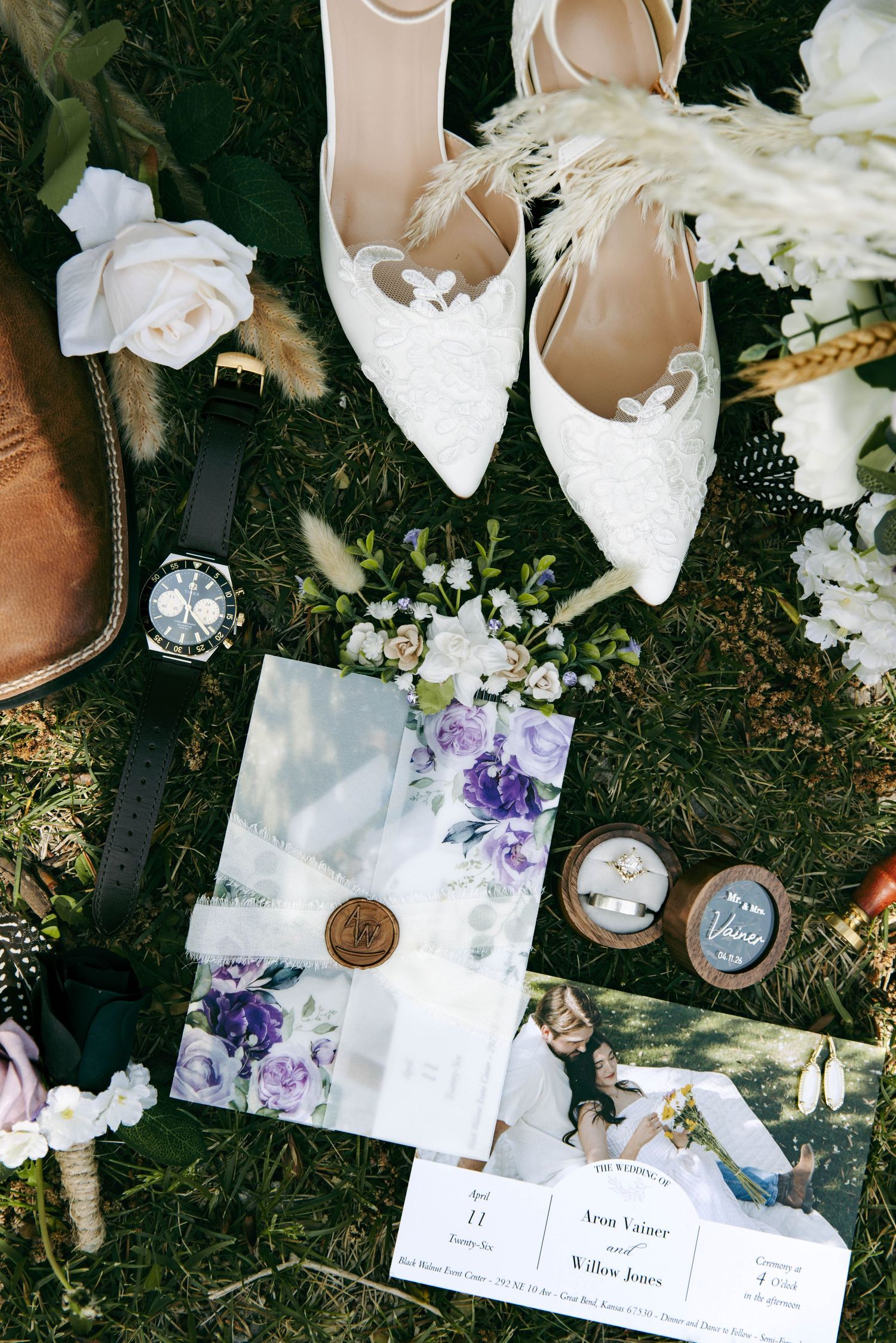 Elegant wedding flat lay featuring lace bridal shoes, floral invitations, rings, a watch, and white flowers on green grass.