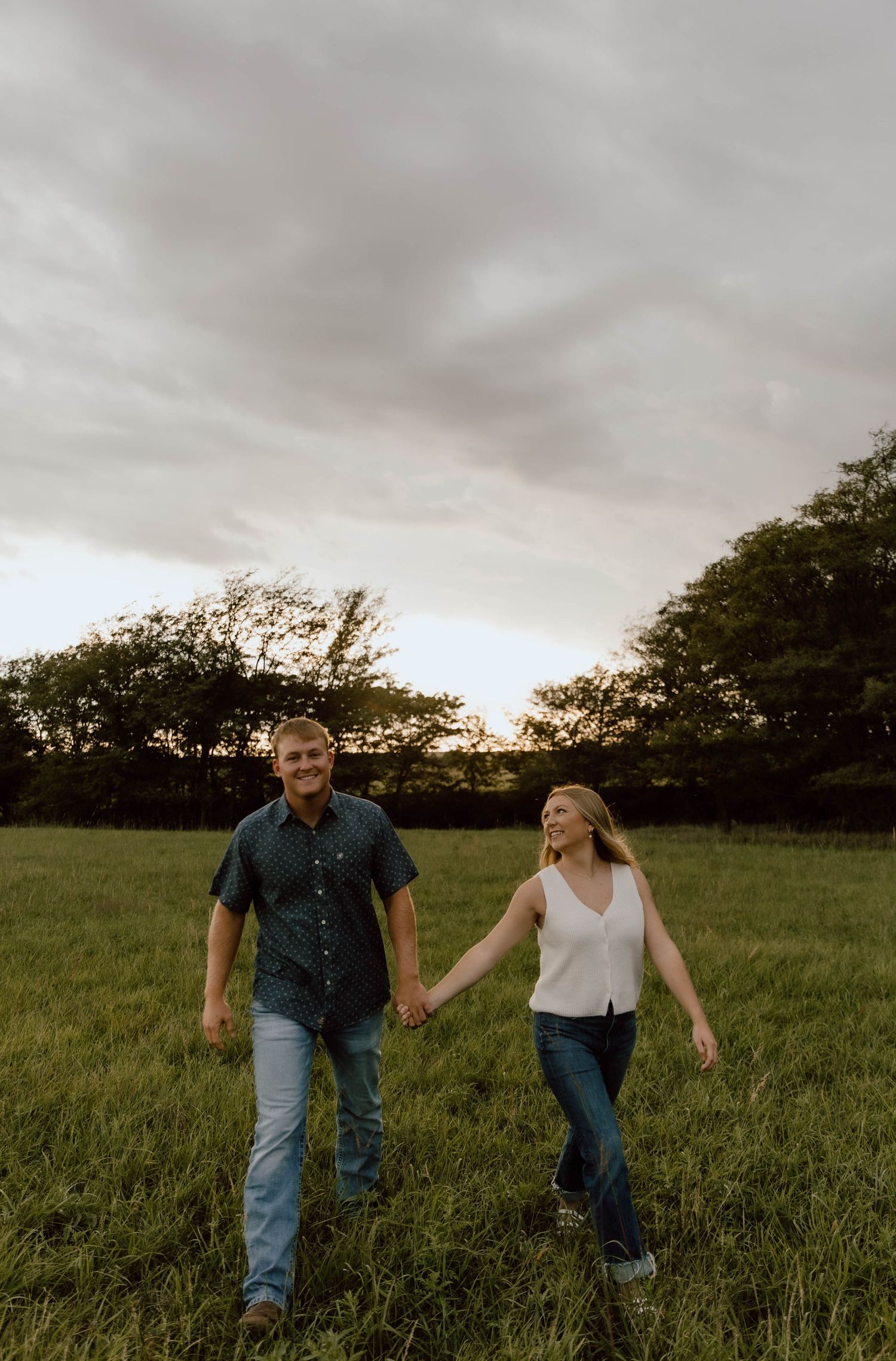 A couple holds hands while walking through a lush green field at sunset under moody clouds.