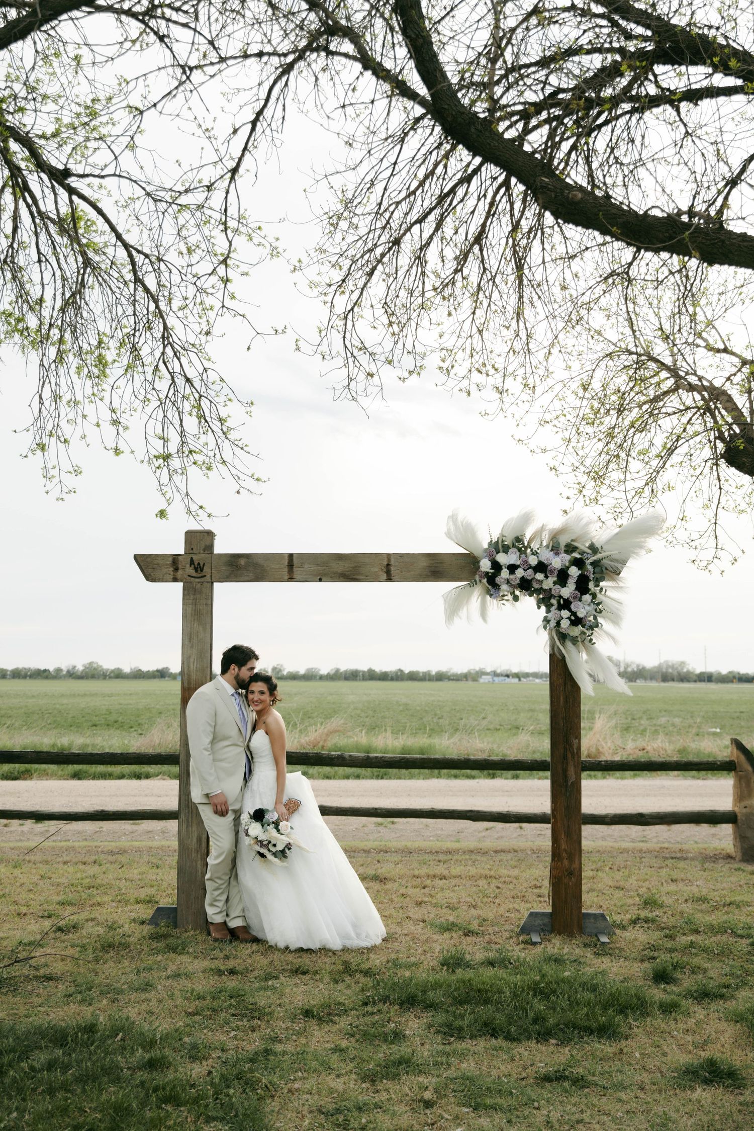 Bride and groom posing under rustic wooden arch with floral decor in open field during outdoor wedding ceremony.