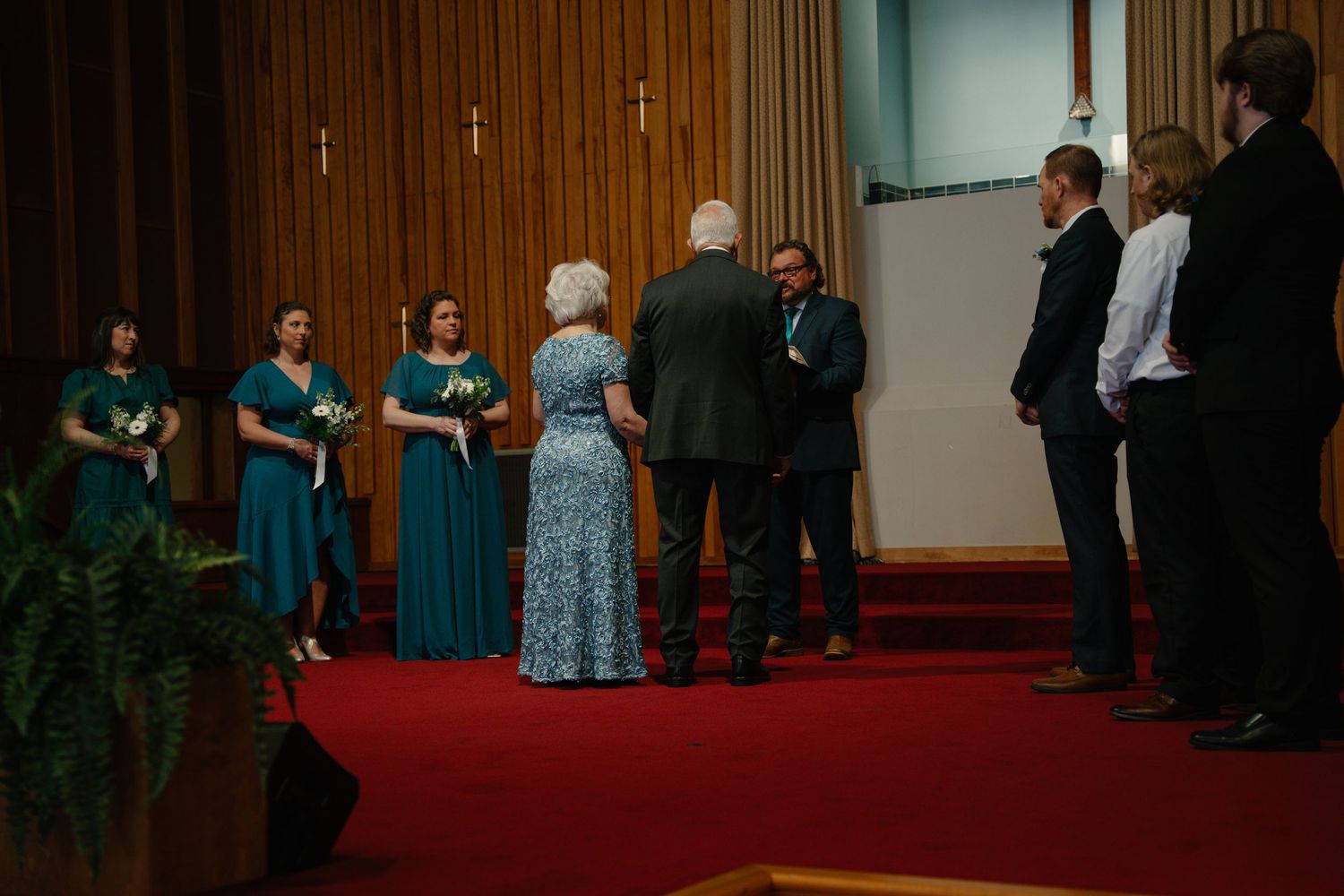 Wedding ceremony in a church with bridesmaids in teal dresses standing on a red carpet aisle.