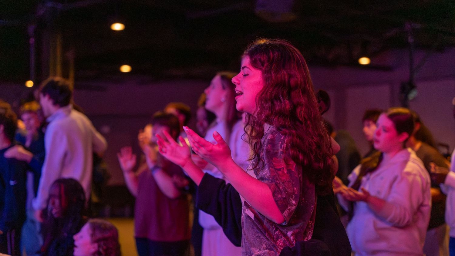Woman with curly hair clapping and singing during a colorful worship service under pink and blue lights. in Abingdon Maryland