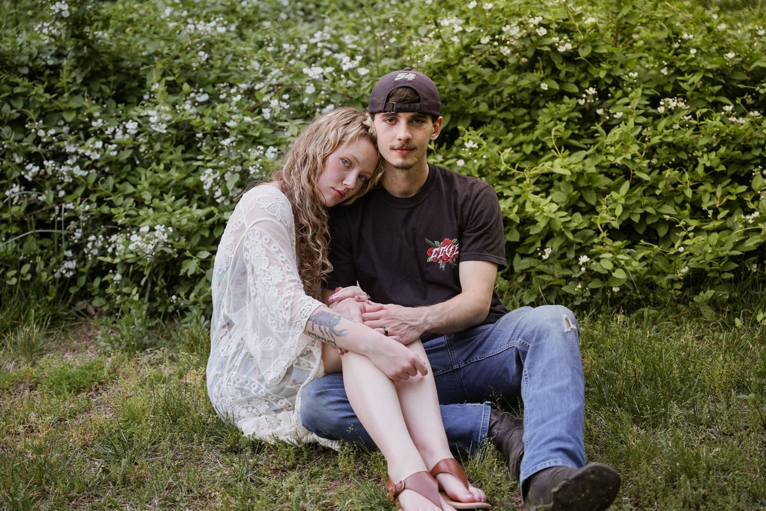 A young couple sits together on green grass, leaning close, surrounded by lush white flowering bushes outdoors. In Joppatowne Maryland