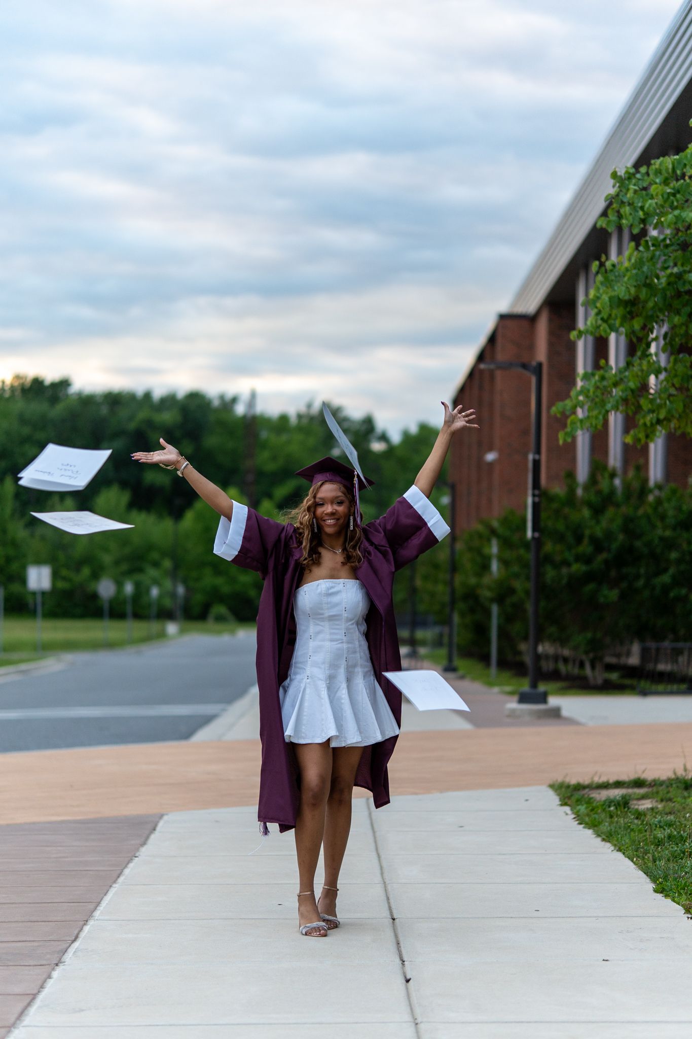 A smiling graduate in maroon cap and gown tosses papers in the air while walking on a campus sidewalk. In Havre de grace