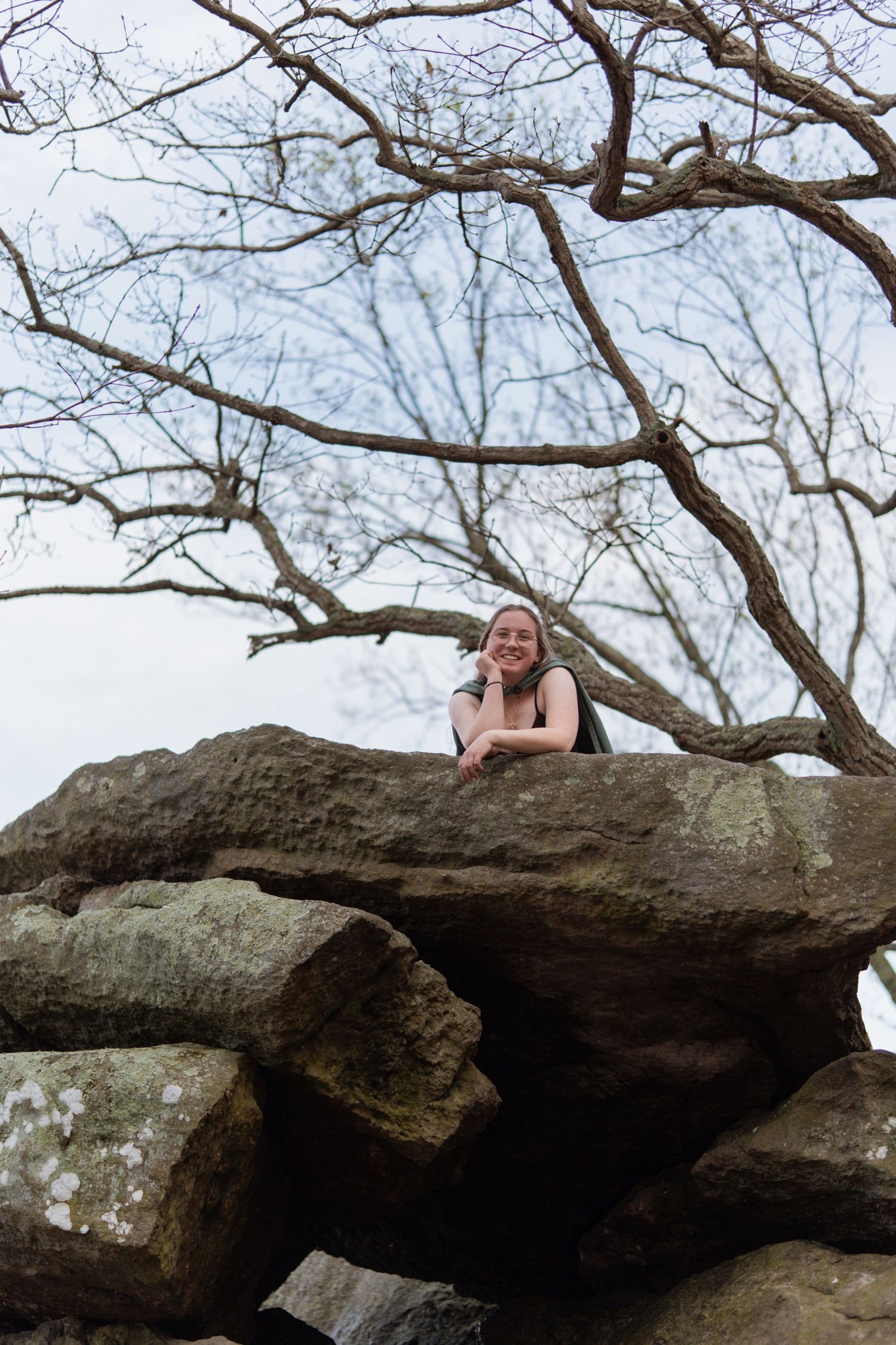 Young woman leaning over large rocks, smiling, with bare tree branches against sky. in Maryland Rock state park Senior