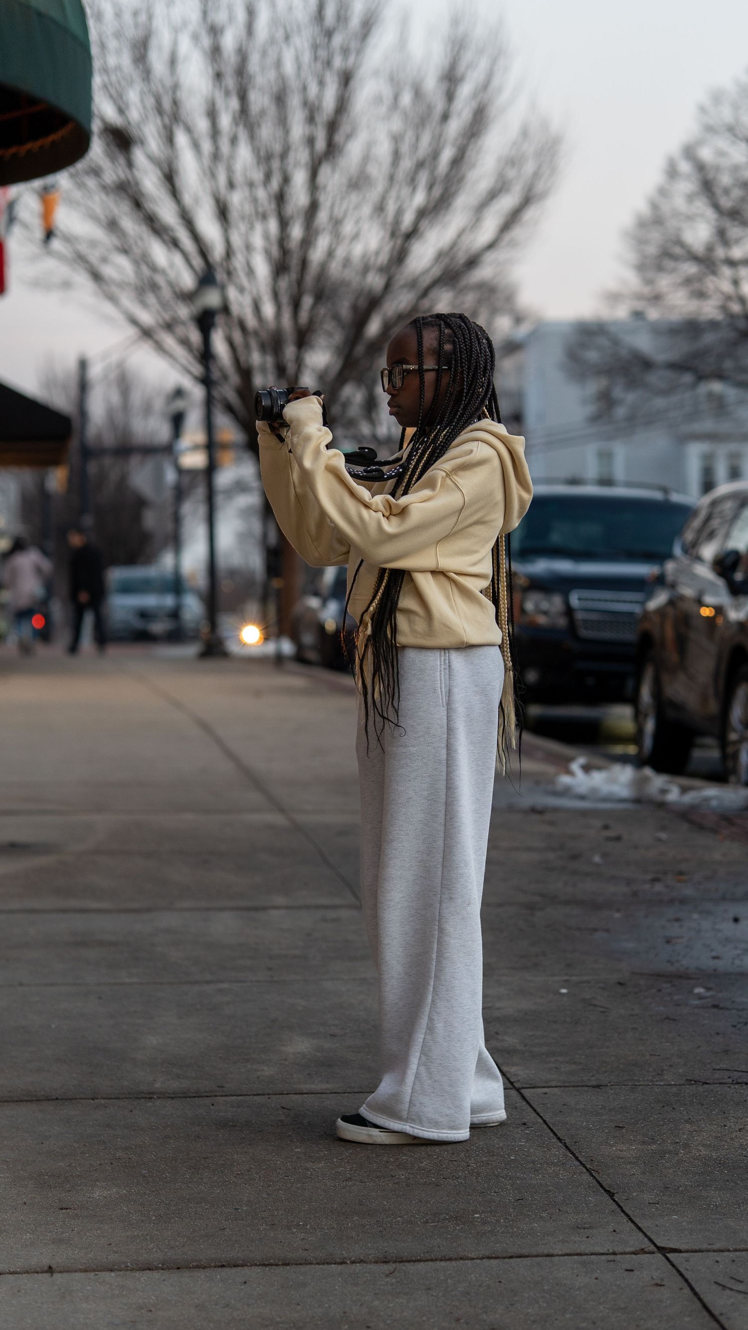 Young woman with braids in cream hoodie and wide-leg pants photographs outdoors on urban sidewalk.In Bel Air Maryland