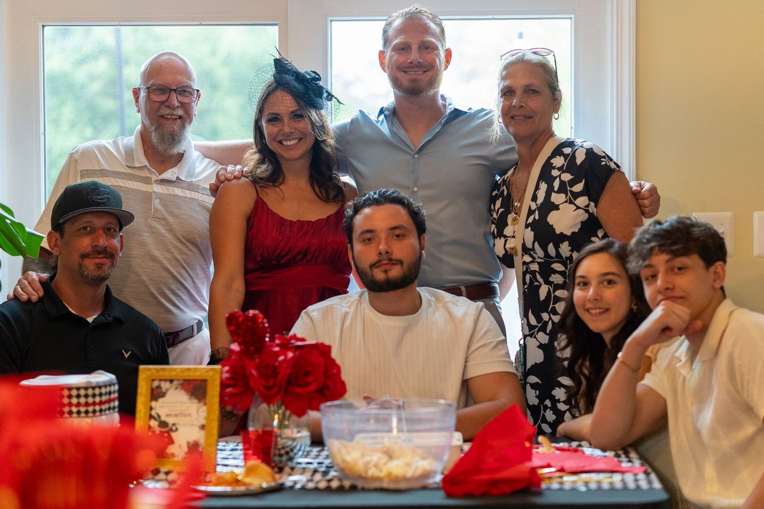 Group of eight adults posing together at a festive party with red floral decorations on table. In Forest Hill Maryland