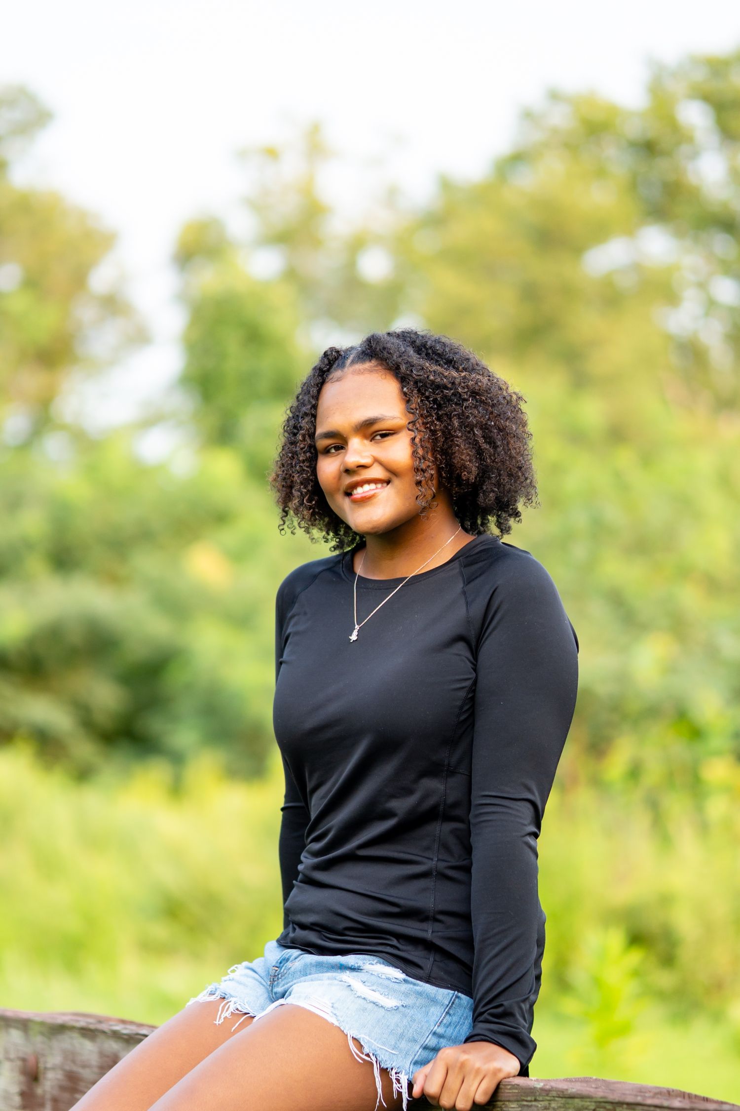 Young Black woman with curly hair smiling outdoors, wearing black long-sleeve top and denim shorts.