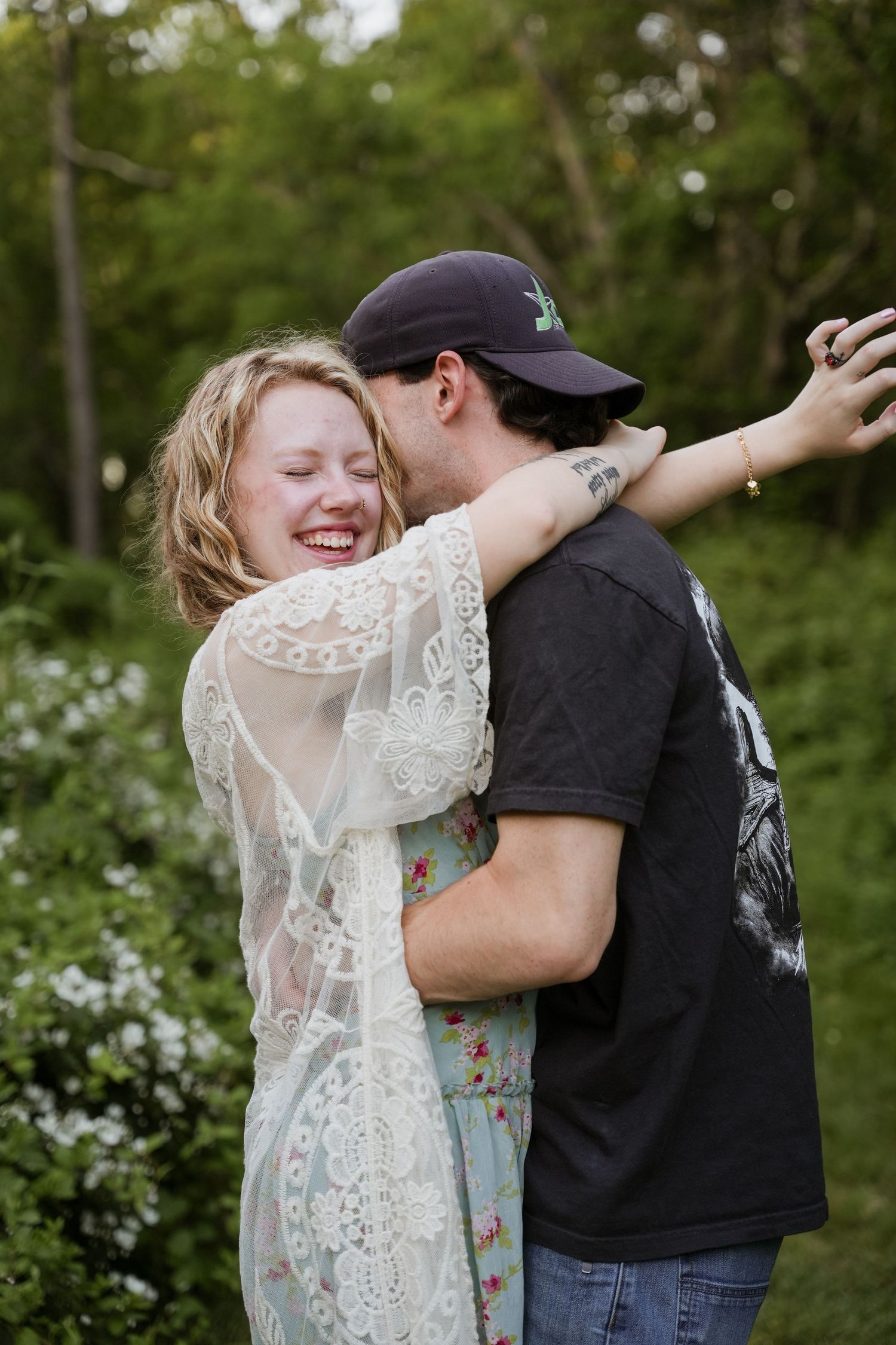 A laughing woman in lace embraces a man in a black cap outdoors among green trees and white flowers.