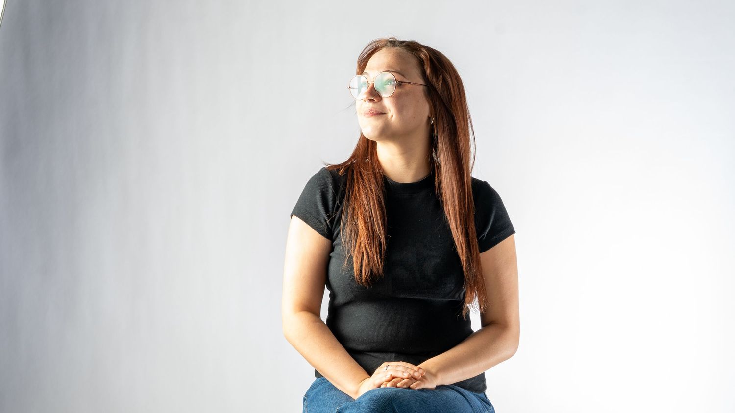 Young woman with long red hair and glasses sitting confidently in a black t-shirt and jeans against white background. In Bel Air Maryland