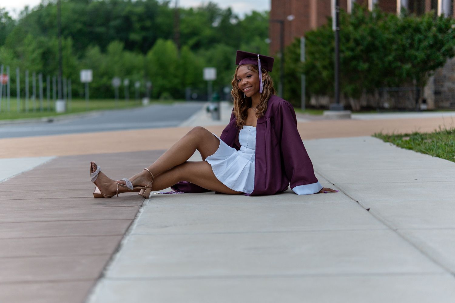 A young woman in a maroon graduation gown and white dress sits relaxed on a campus sidewalk smiling. In Havre de grace