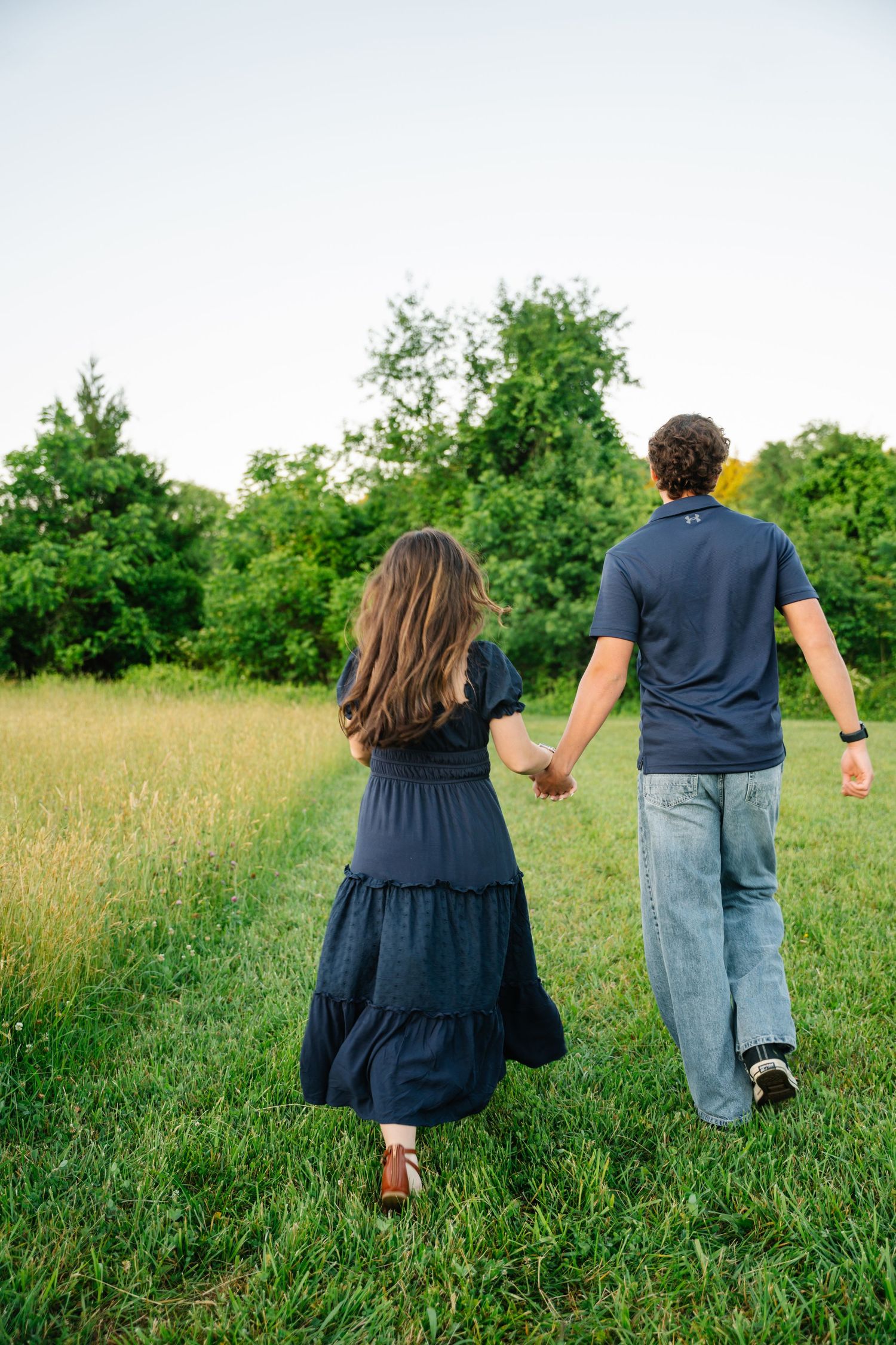 A couple holding hands walking through a lush green field, woman in navy dress and man in blue shirt and jeans.