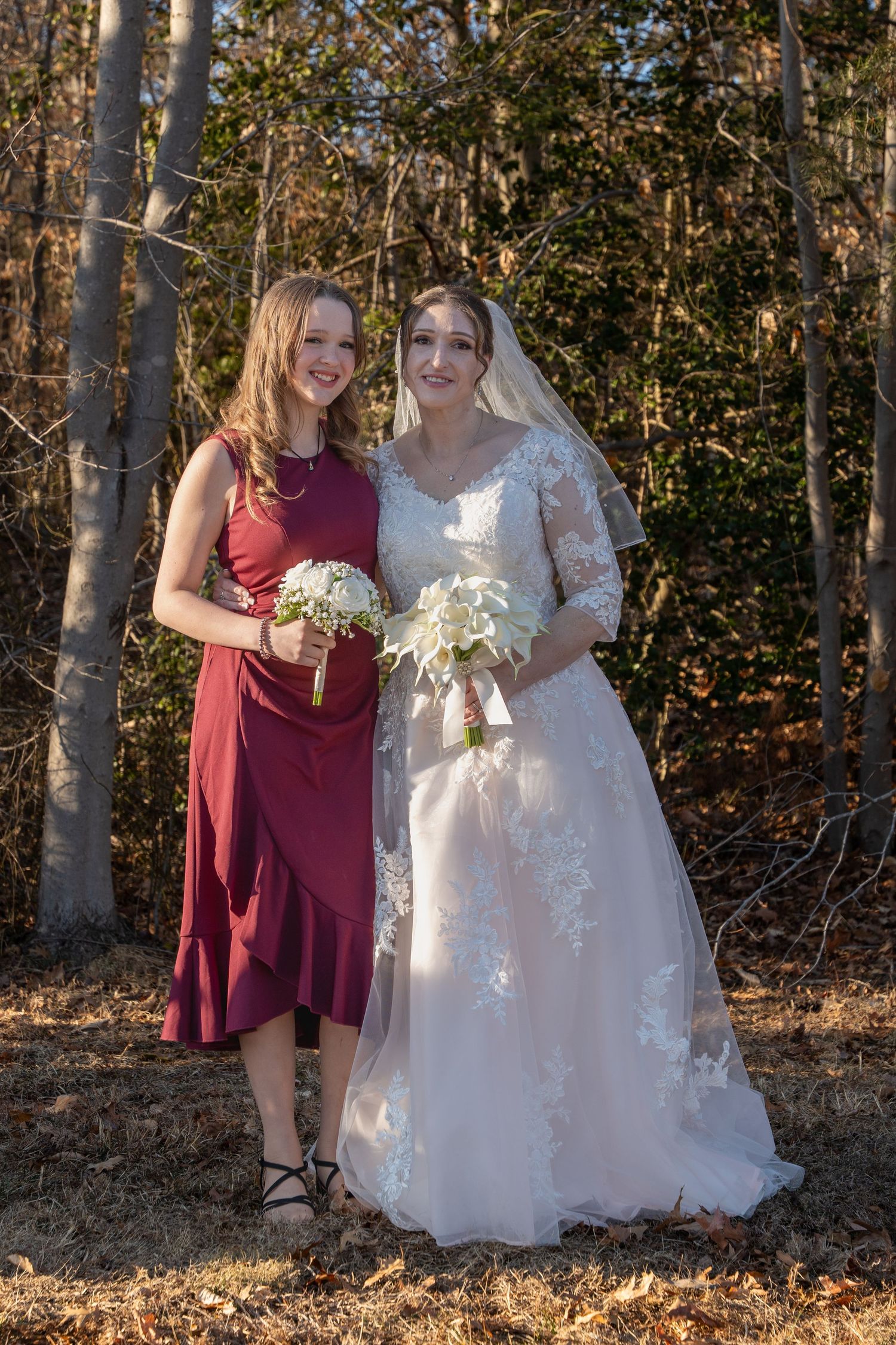Bride in lace wedding gown poses with bridesmaid in burgundy dress holding bouquets in autumn woodland.in Abingdon Maryland