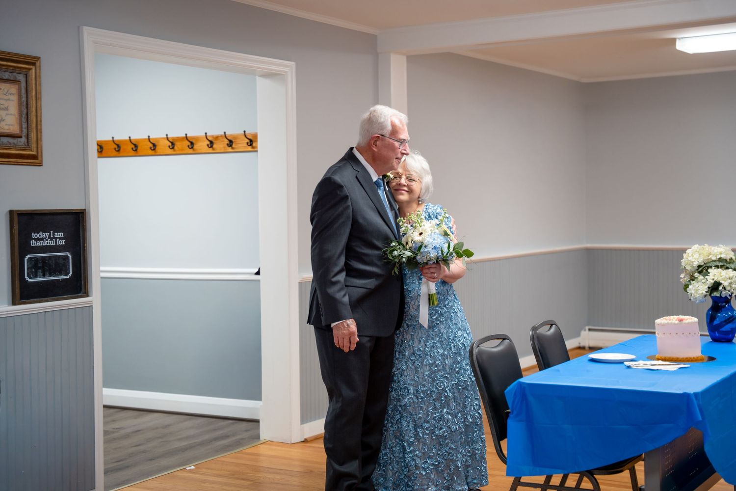 Elderly couple embracing at a wedding reception, woman holding blue bouquet, blue tablecloth visible in background. In Bel Air Maryland