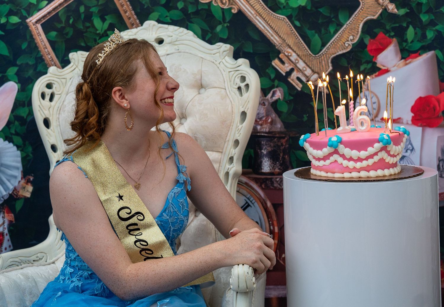 A teenage girl in a blue dress and Sweet 16 sash sits beside a birthday cake with candles at her celebration. In Forest Hill Maryland