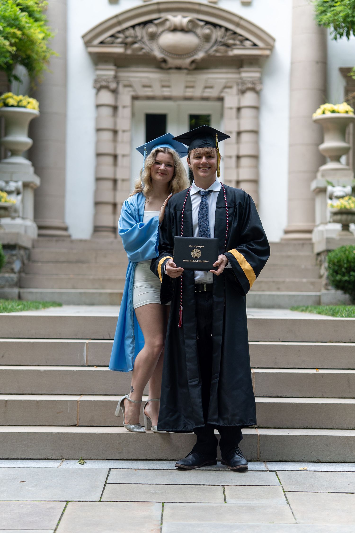 Two graduates in cap and gown posing on steps, one holding a diploma, celebrating their graduation day. In Bel Air Maryland