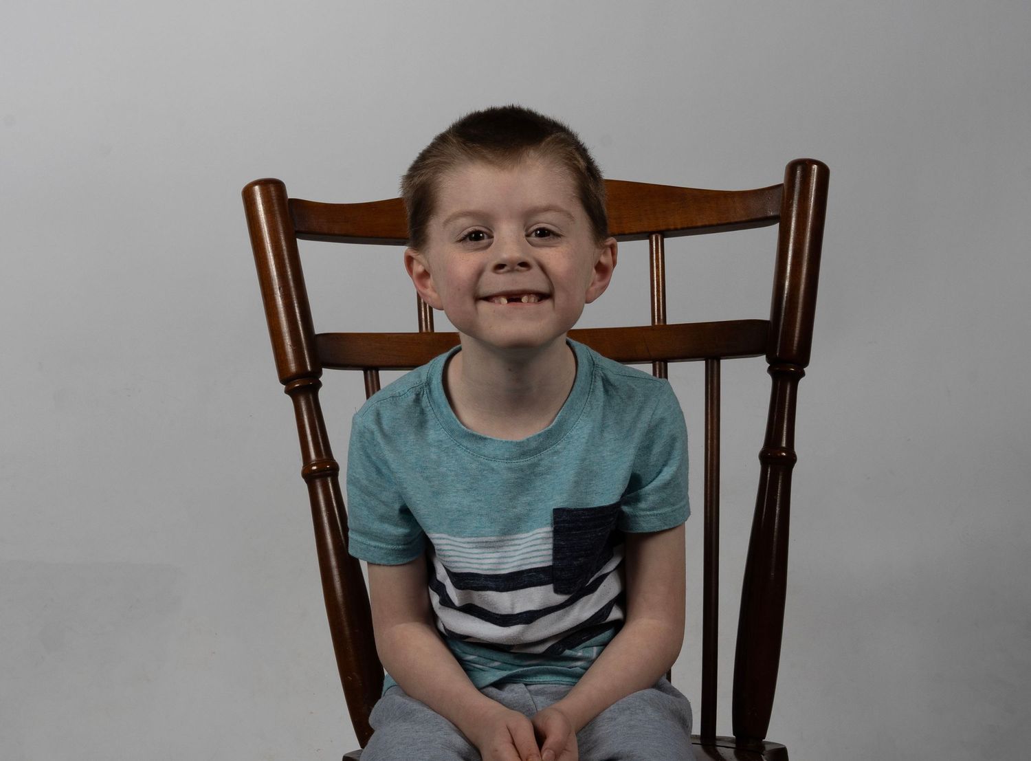 A happy young boy with a missing tooth sits smiling in a wooden rocking chair wearing a striped t-shirt. In Bel Air Maryland