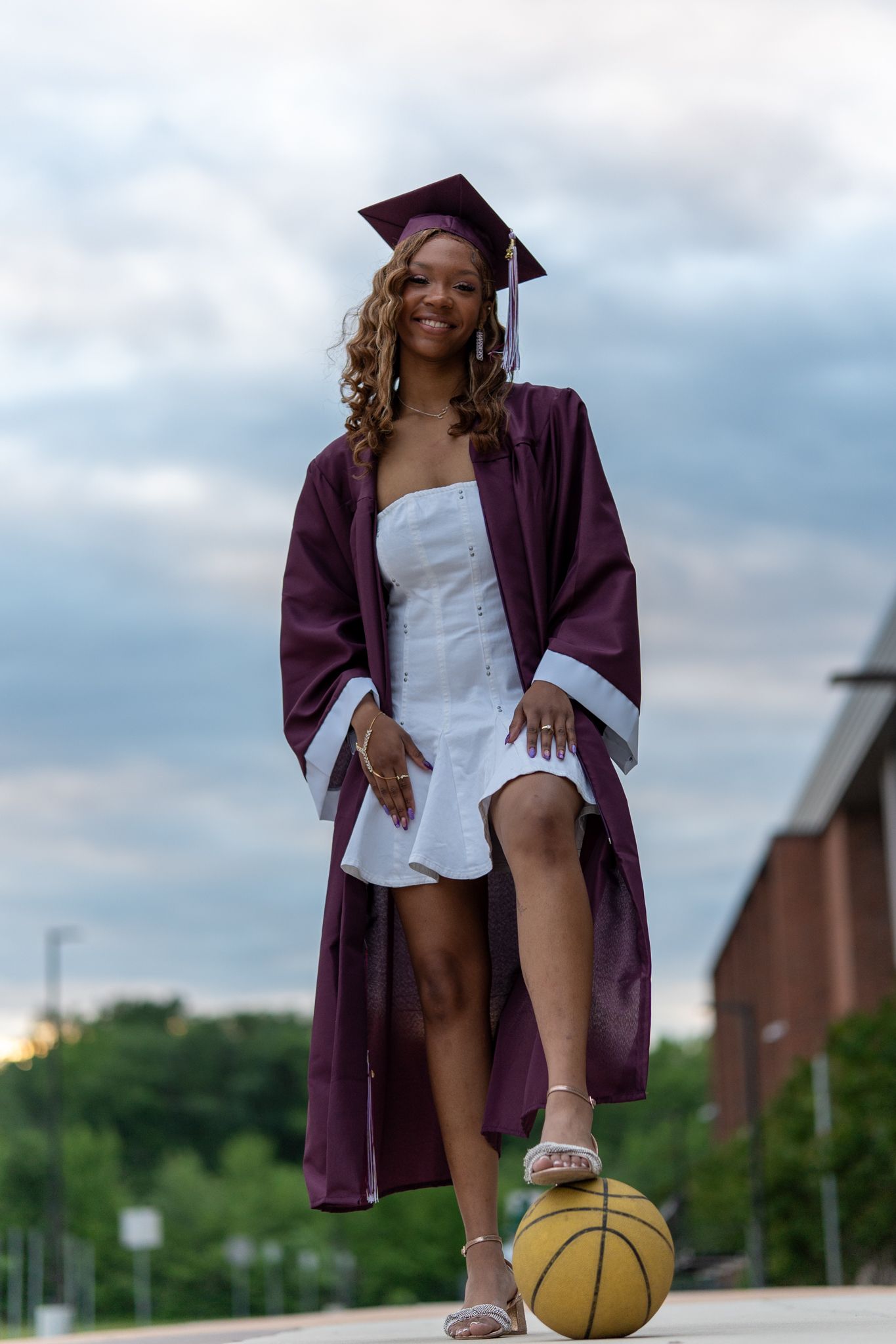 A smiling graduate in maroon cap and gown poses with one foot on a basketball on a campus walkway. In Havre de grace