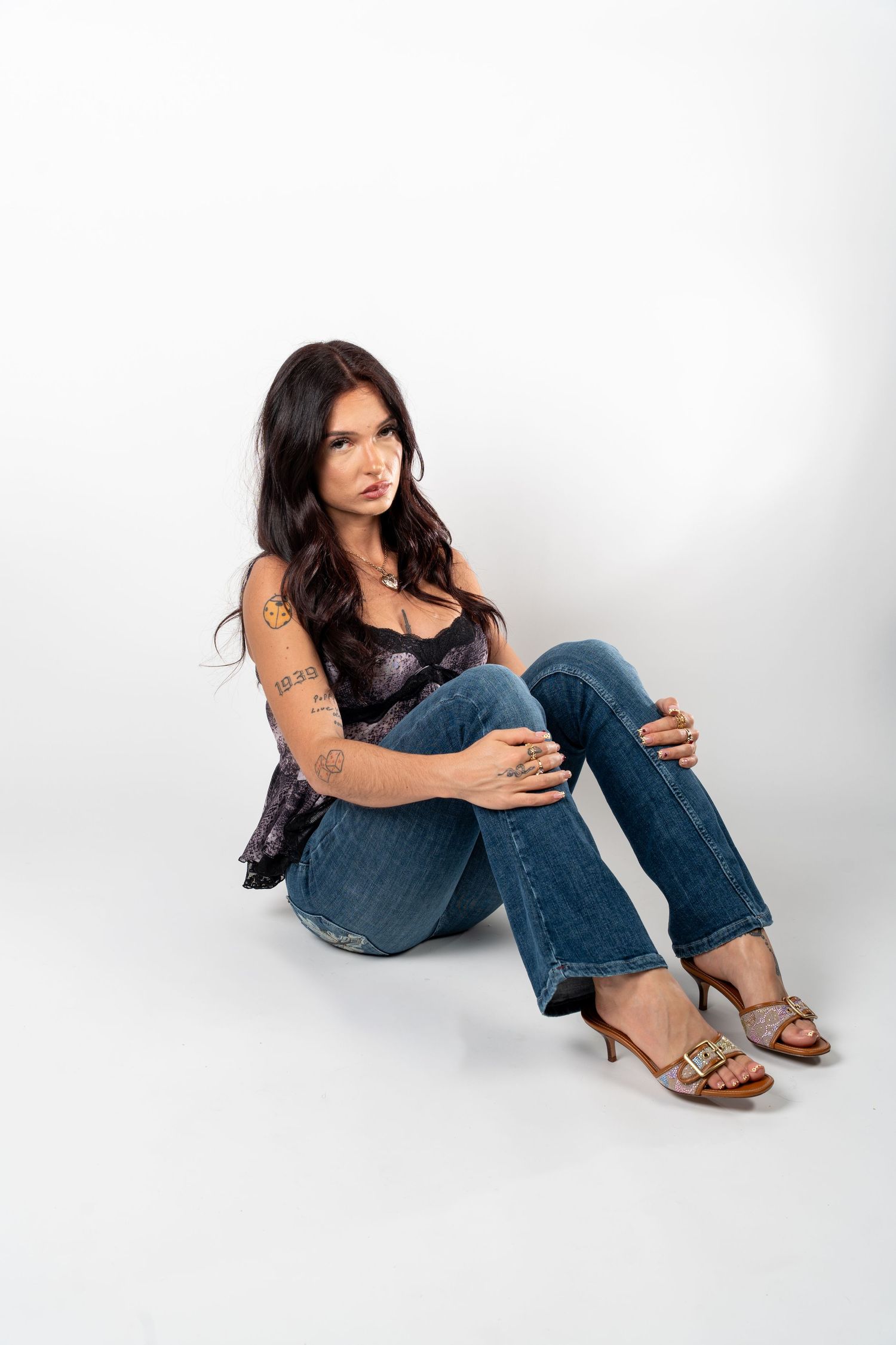 Tattooed brunette woman sits on floor in jeans and heeled sandals posing against a white studio background. In Bel Air Maryland