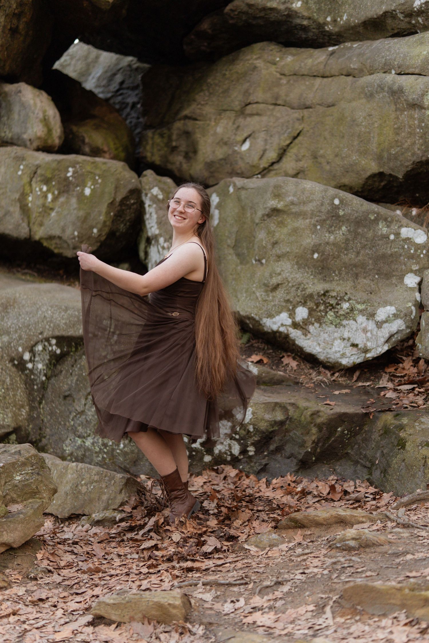 Smiling young woman in brown dress holding skirt, posing among large mossy rock formations. in Maryland Rock state park Senior