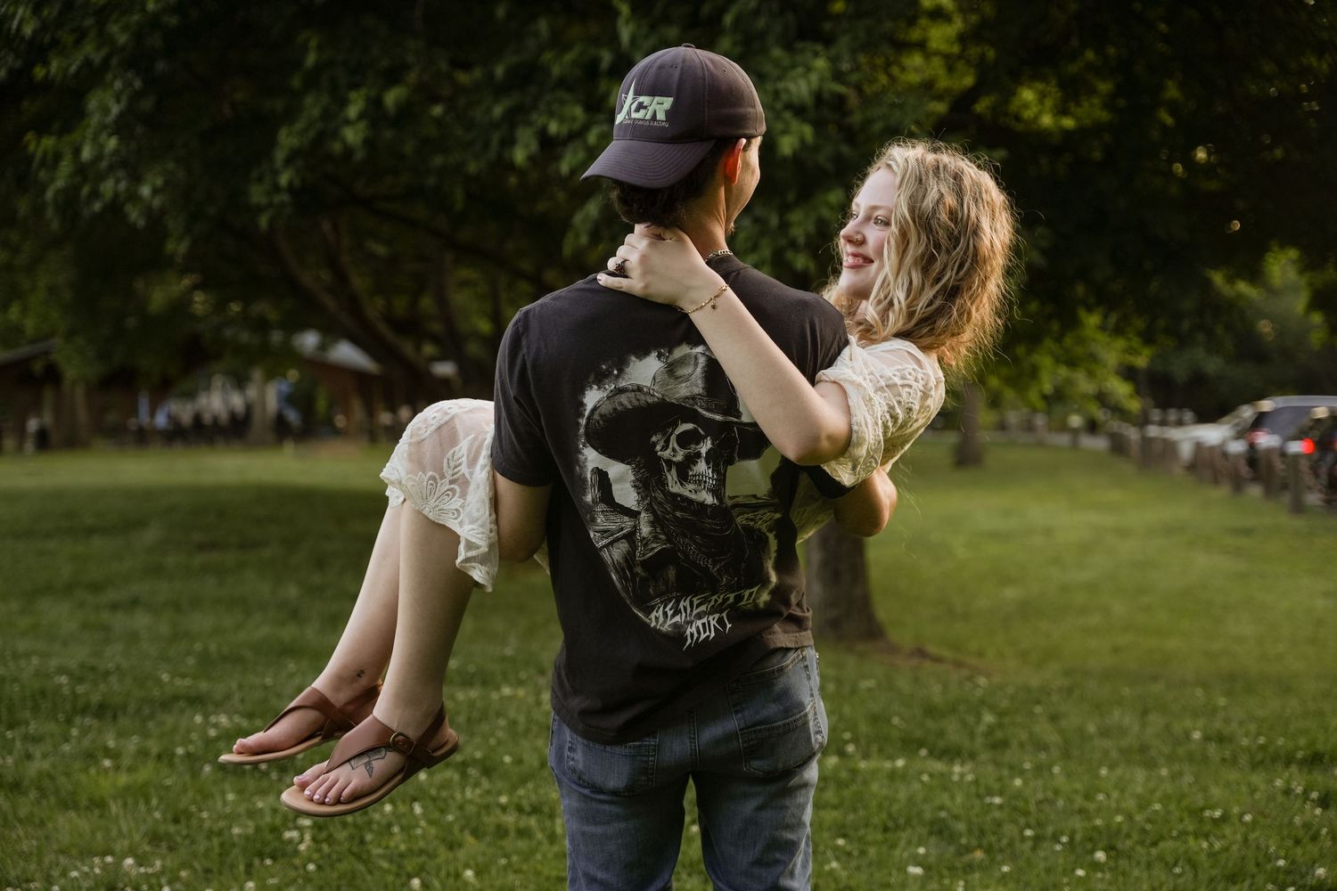 A man lifts a laughing woman in a grassy park, both wearing casual T-shirts on a warm summer evening. In Joppatown Maryland