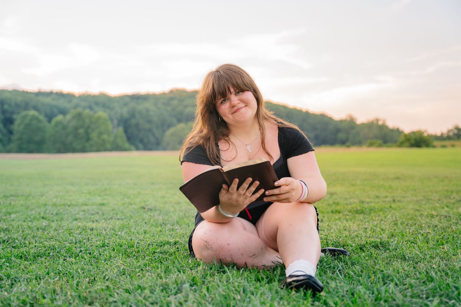 Young woman sitting on green grass in a field, reading a book at sunset with trees in the background.