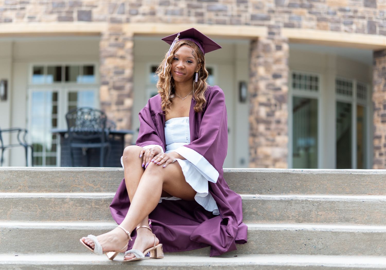 Young woman in maroon graduation cap and gown sitting on stone steps, celebrating her academic achievement. In Havre de Grace Maryland