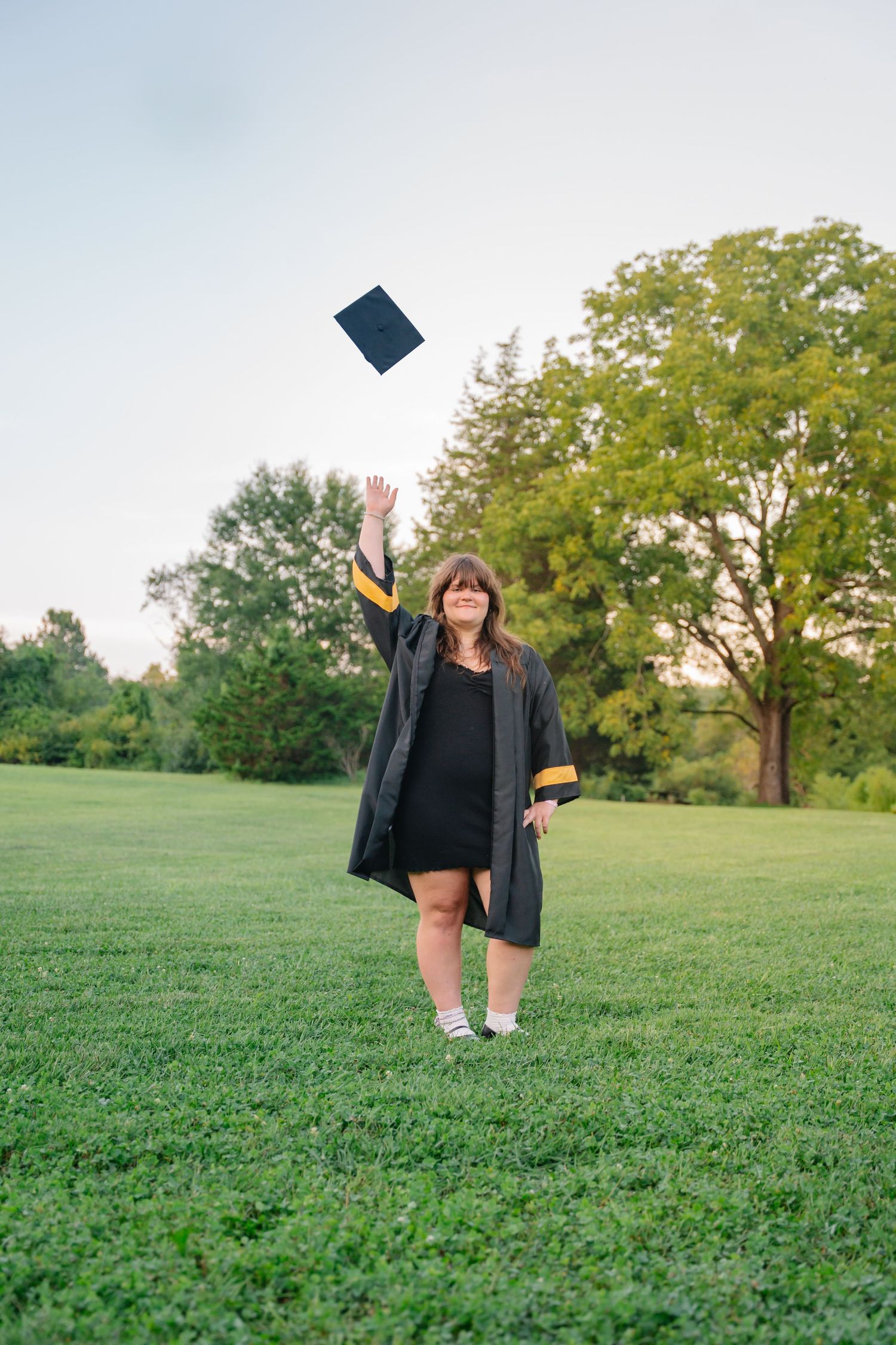 Woman in graduation gown tossing cap in the air on green lawn with trees in background.