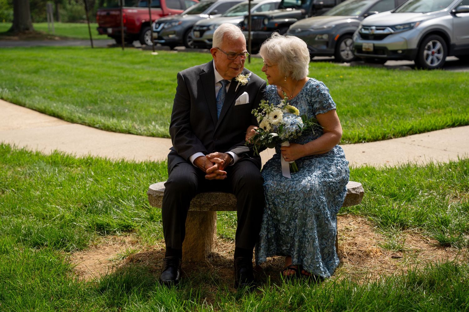 Elderly couple in formal attire sitting on a stone bench in a park, sharing a tender moment with a bouquet of white flowers. In Bel Air MD