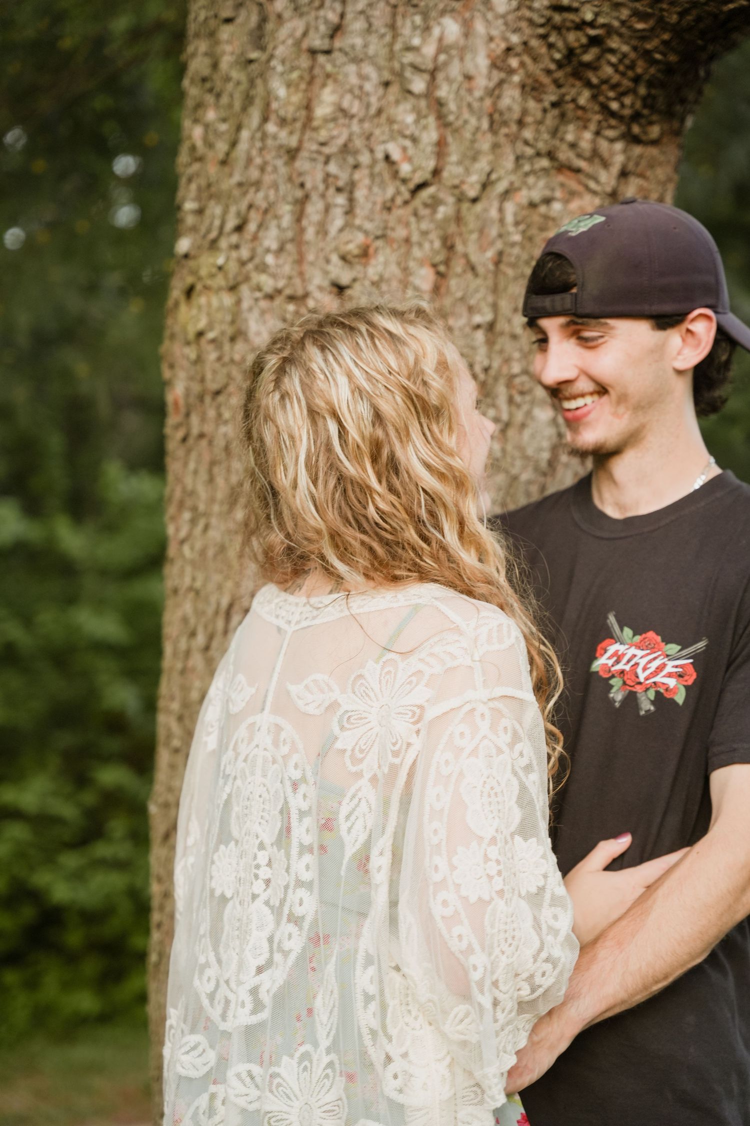 A smiling young couple sharing a tender moment together in front of a large tree in a lush green forest. JoppaTowne Maryland