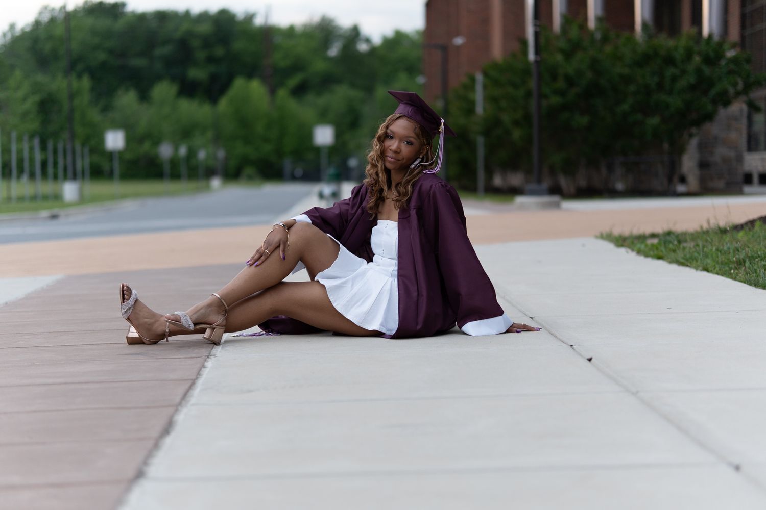 A graduate in a maroon cap and gown sits casually on a sidewalk near a brick building smiling softly. In Havre de grace