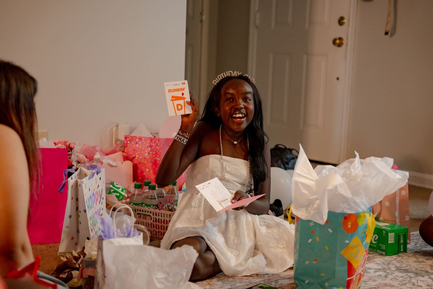 Bel Air Maryland, Young woman wearing a tiara opens birthday gifts surrounded by colorful gift bags while smiling joyfully.
