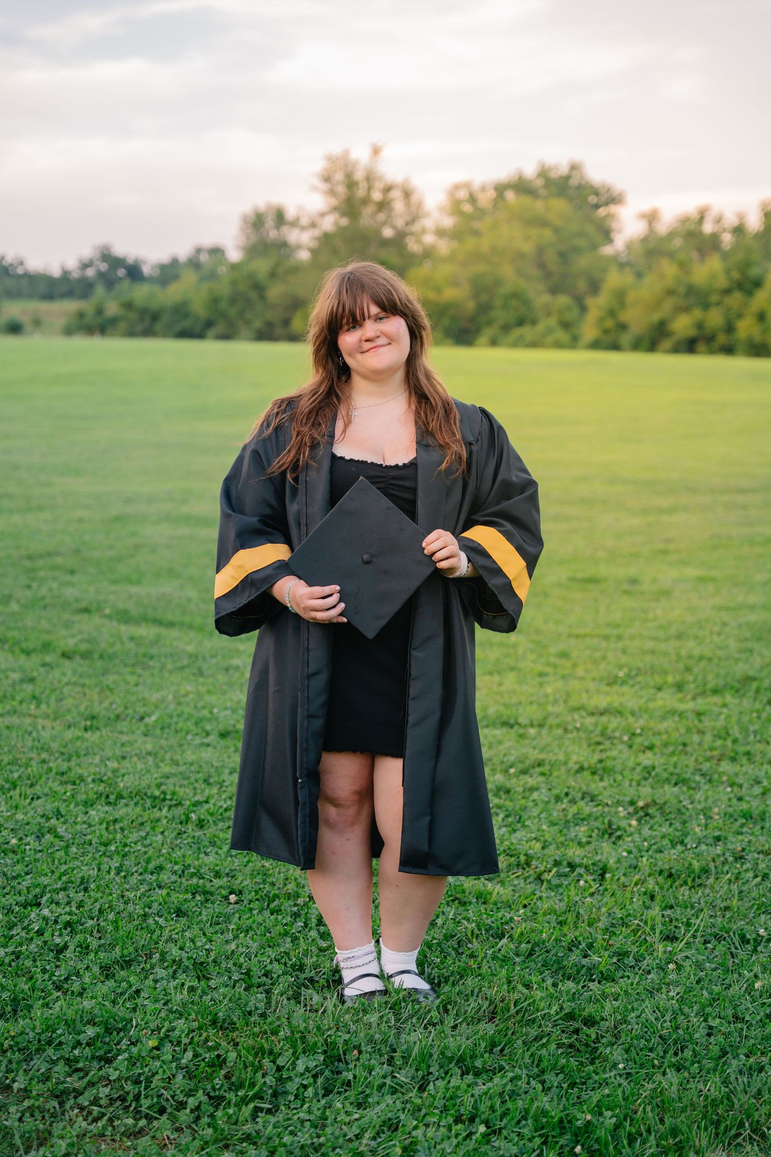 Young woman in black and gold graduation gown holding cap while standing on green grass field at sunset.