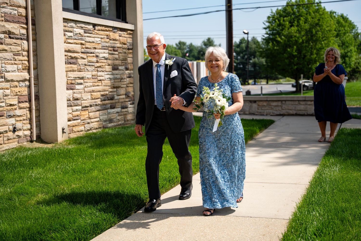 Elegant couple walking together on a sidewalk, woman in blue lace dress holding white bouquet, man in black suit. In Bel Air Maryland