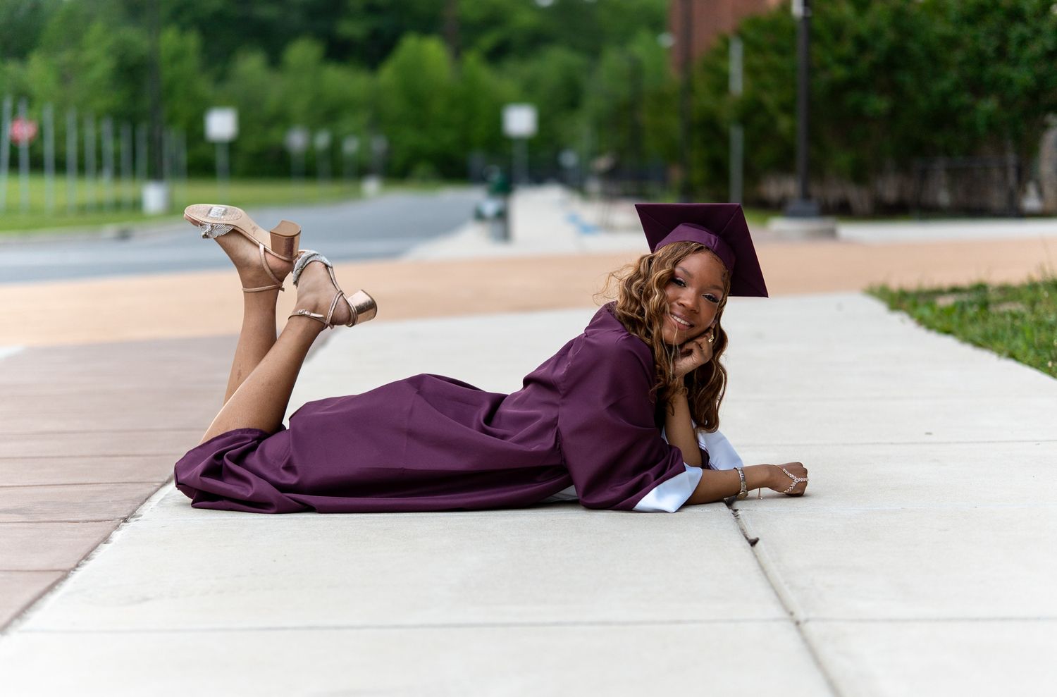 Young woman in maroon graduation cap and gown poses joyfully on a sidewalk outdoors on graduation day.