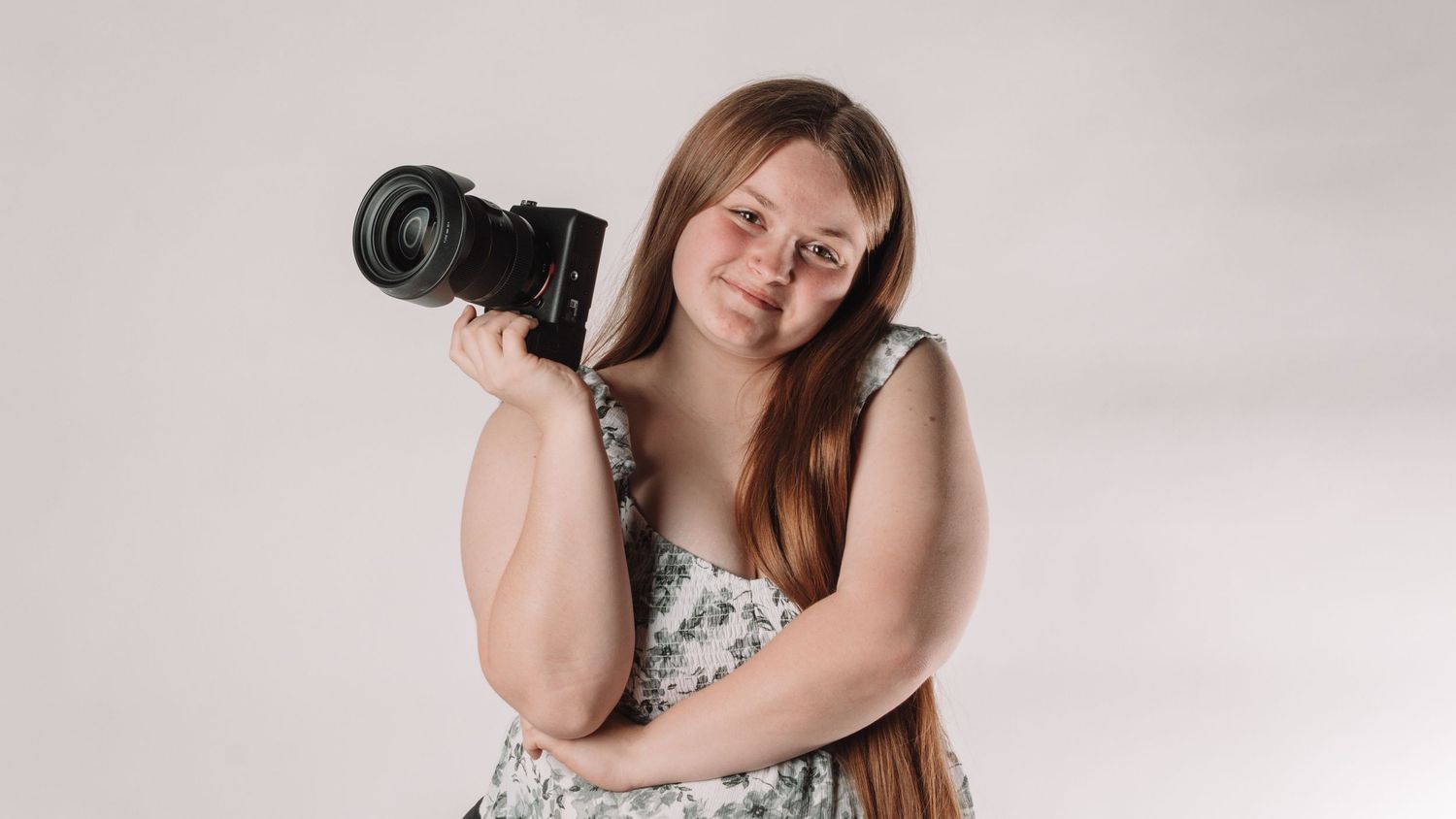 Young woman holding a professional Mirrorless camera over her shoulder, smiling in a floral dress against a white background. in Bel Air MD
