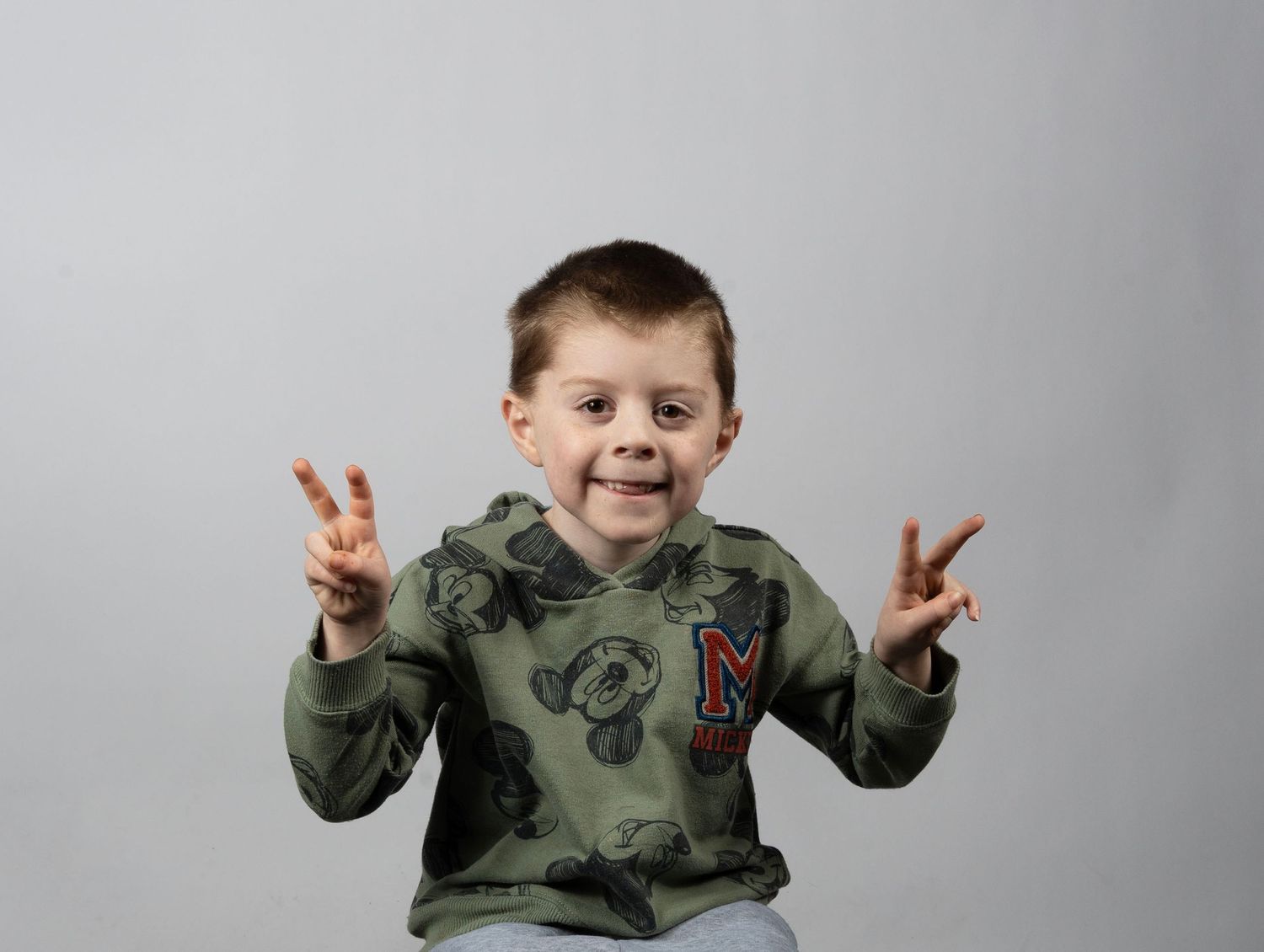 A smiling young boy in a Mickey Mouse hoodie makes peace signs with both hands against a gray background. In Bel Air Maryland