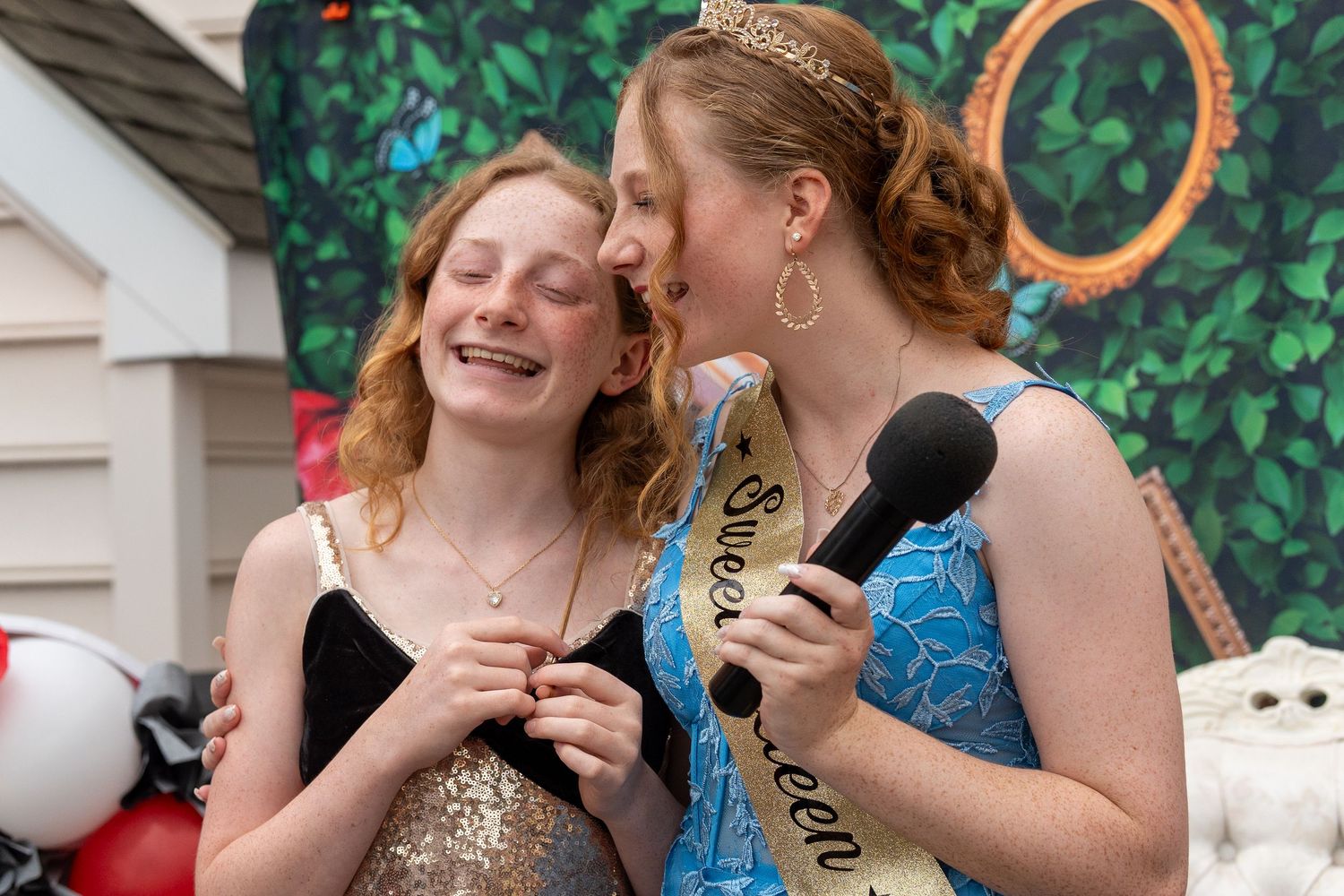 Two teenage girls laugh together, one wearing a Sweet Sixteen sash and tiara holding a microphone. In Forest Hill Maryland