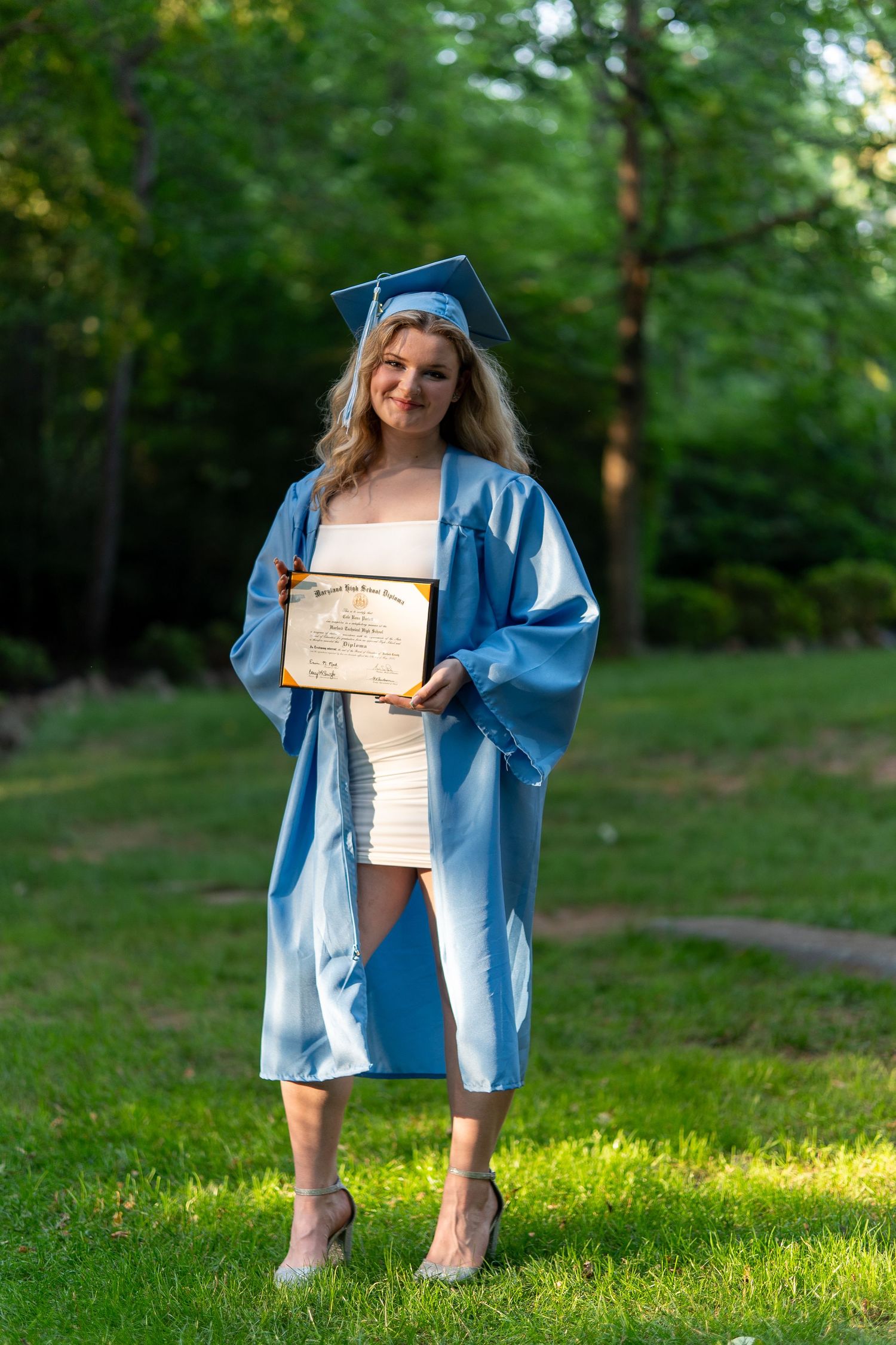 Smiling female graduate in light blue cap and gown proudly displaying her diploma on a sunny lawn.In Bel Air Maryland