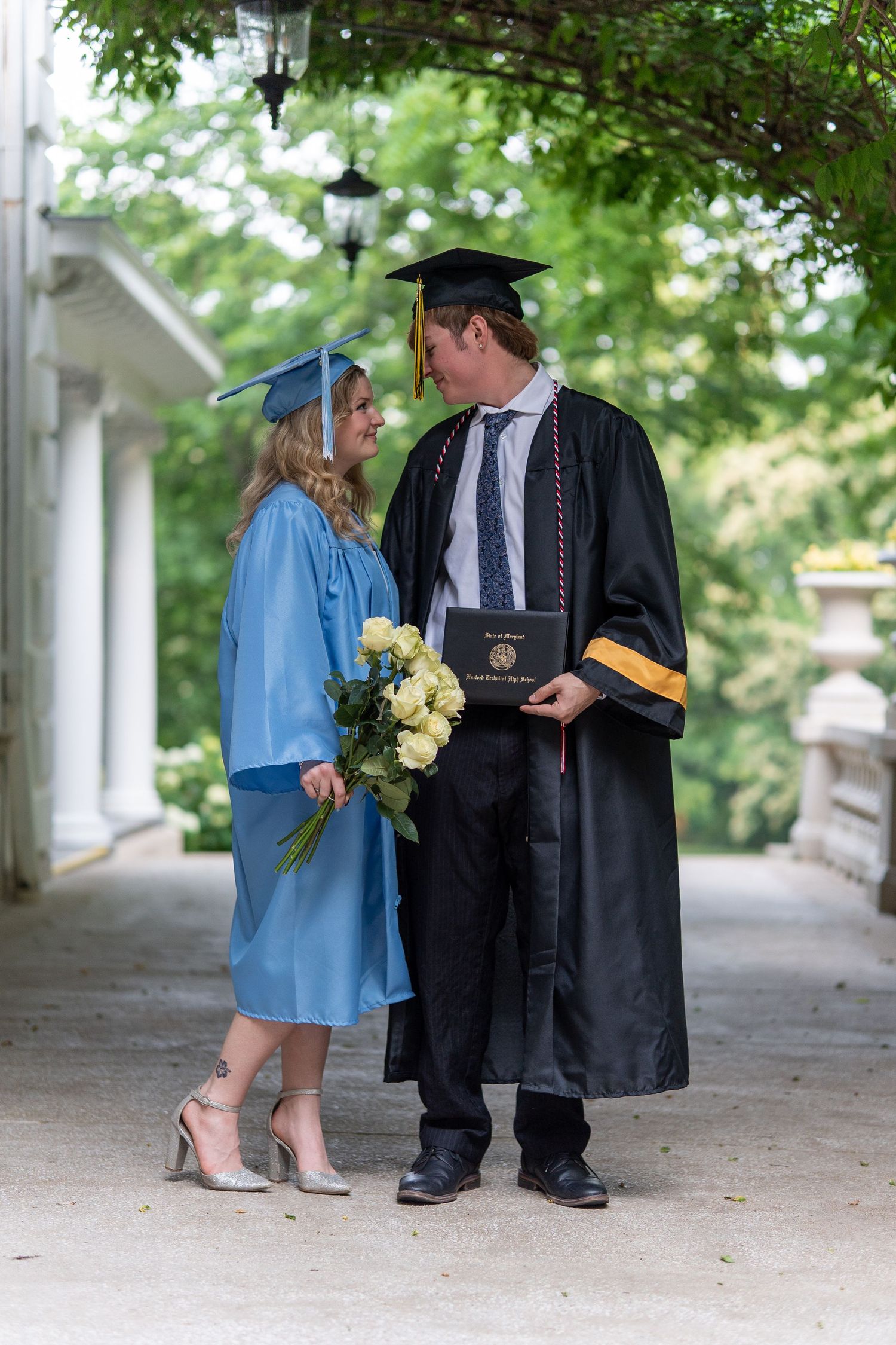 Two graduates in caps and gowns stand face to face, one holding yellow flowers and a diploma, under a garden archway. In Bel Air Maryland