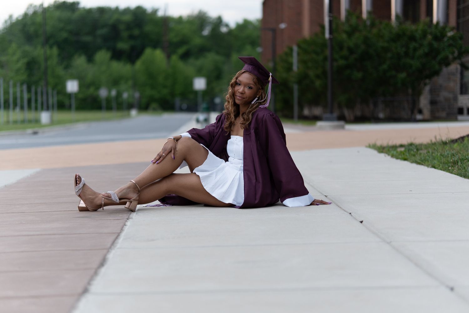 A young Black woman in a white dress and maroon graduation gown sits smiling on a sidewalk outdoors. In Havre de Grace Maryland