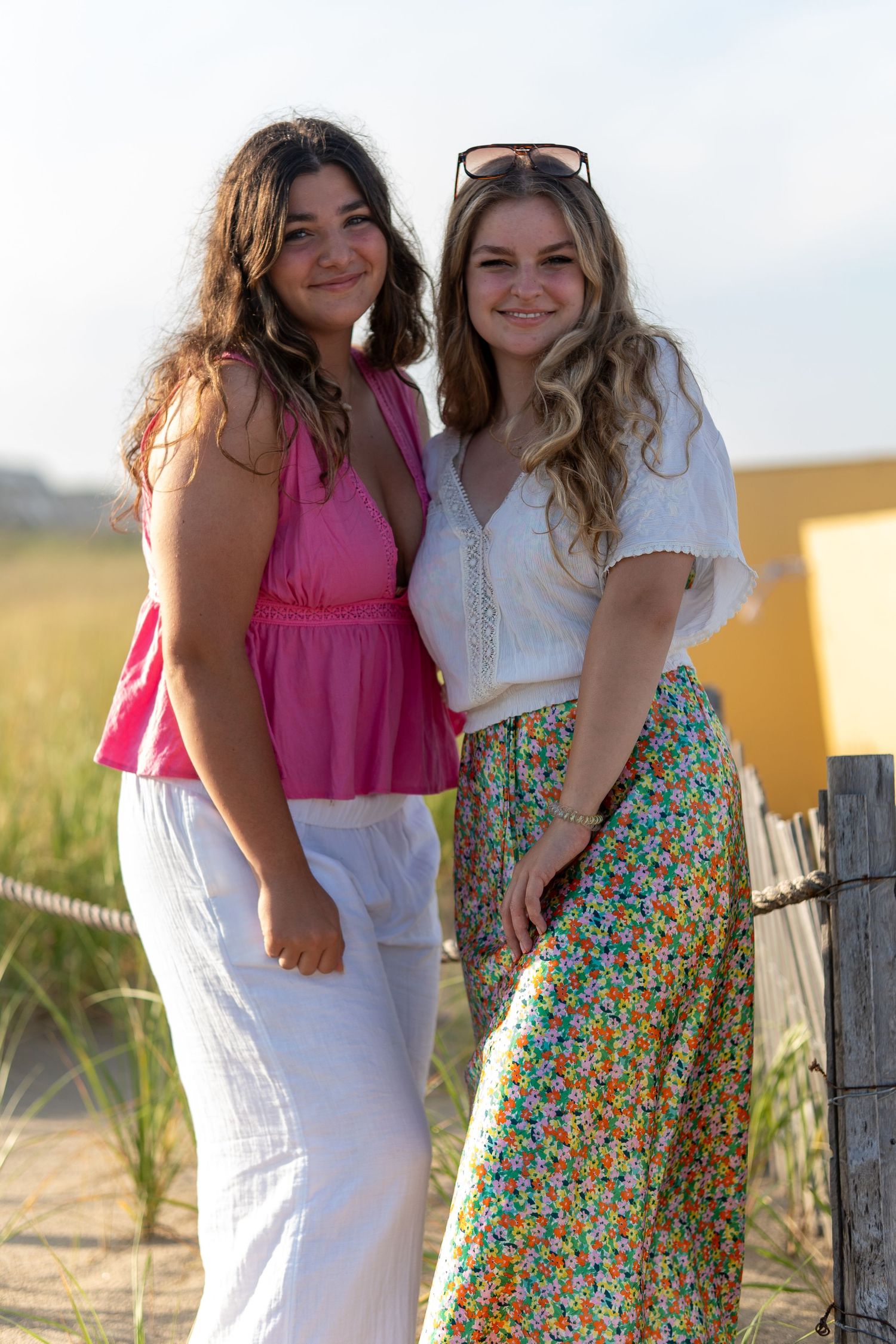 Two young women smile together near a sandy beach fence, one wearing pink and white, the other in a floral skirt. At Bethany Beach