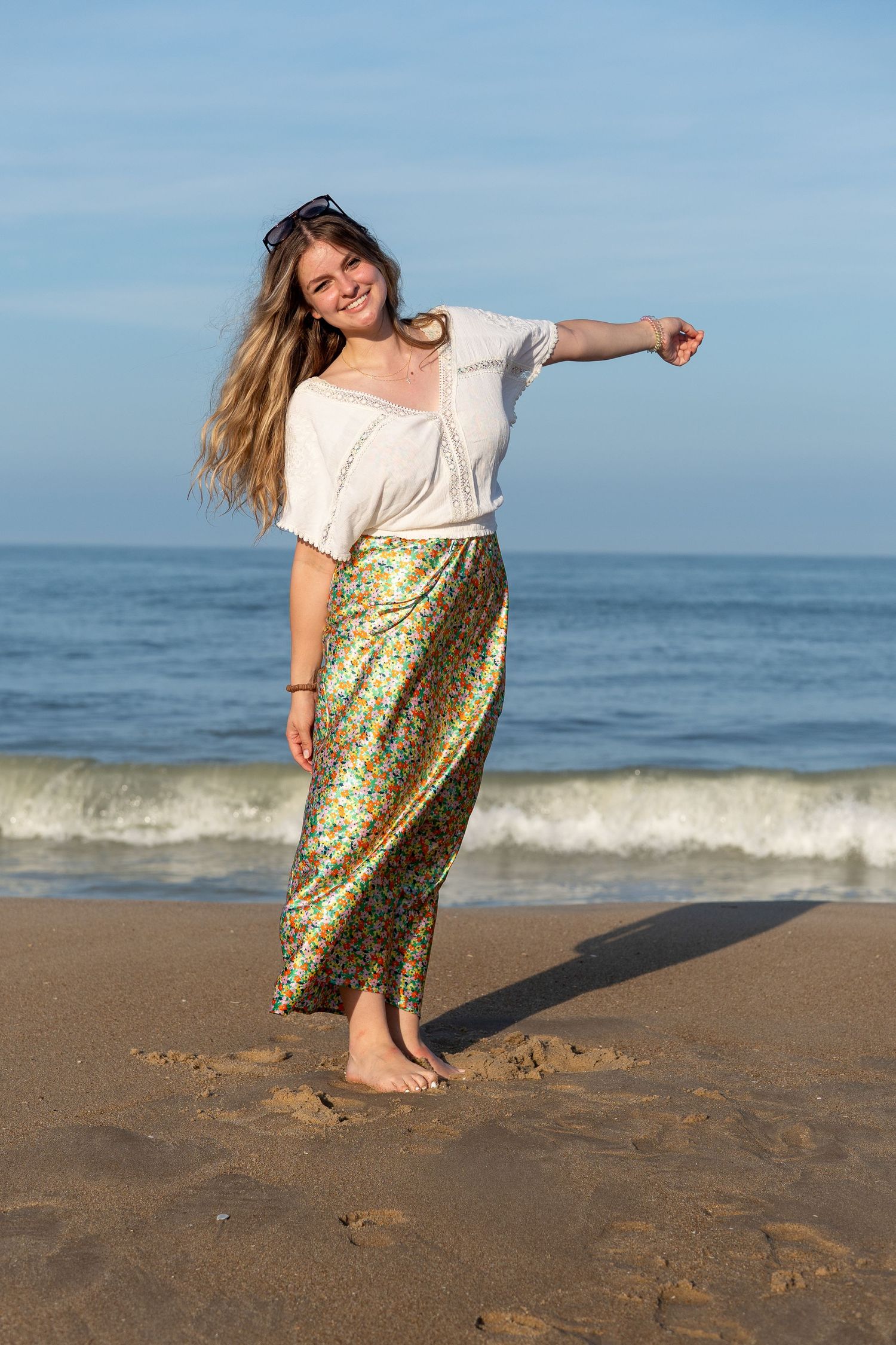 A smiling blonde woman in a white top and colorful floral maxi skirt poses barefoot on a sandy beach at golden hour. At Bethany Beach