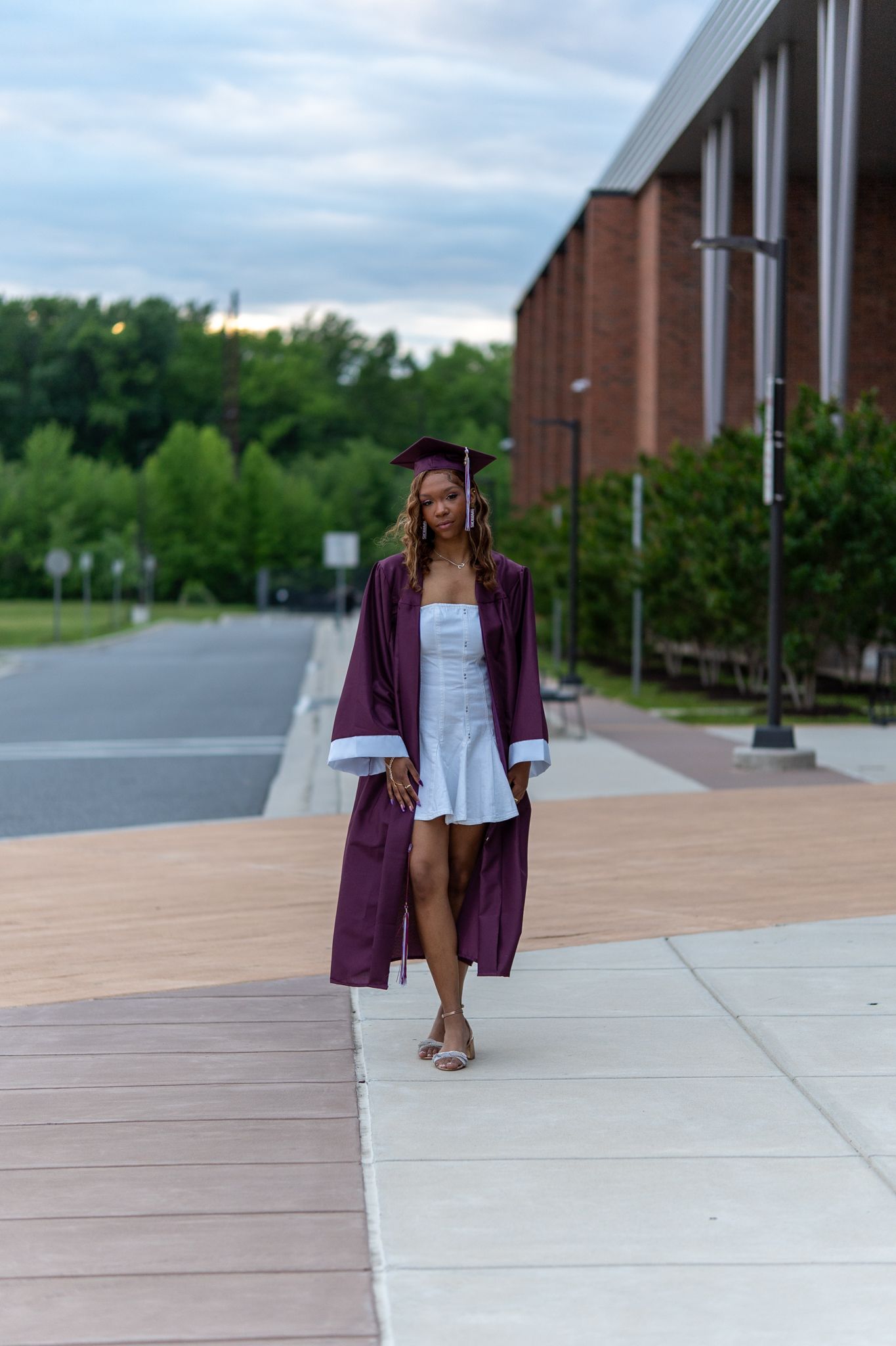 A graduate in a maroon cap and gown walks confidently along a campus sidewalk beside a brick building. In Havre de grace