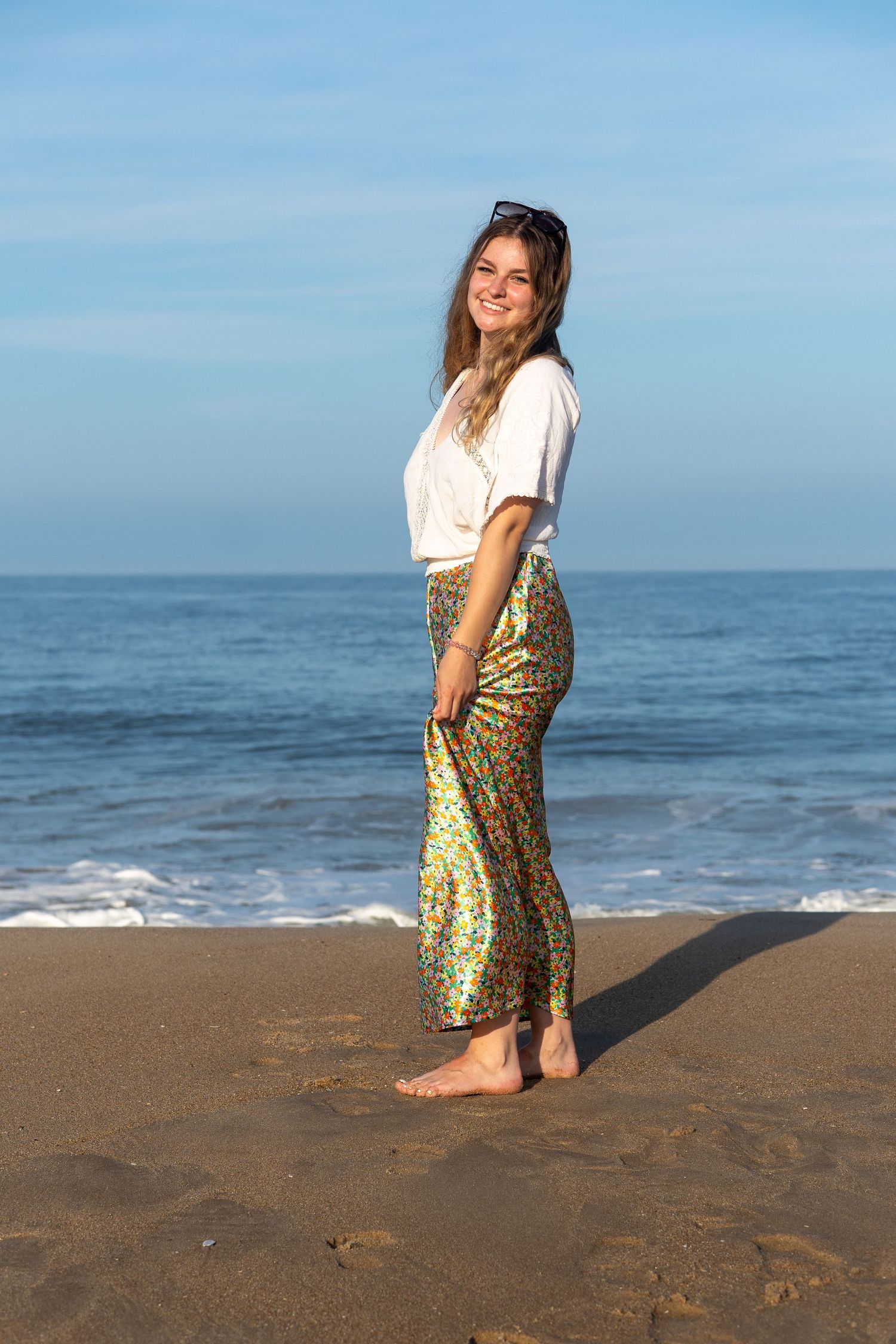 A smiling blonde woman in a white top and colorful floral maxi skirt stands barefoot on a sandy beach at sunset. At Bethany Beach